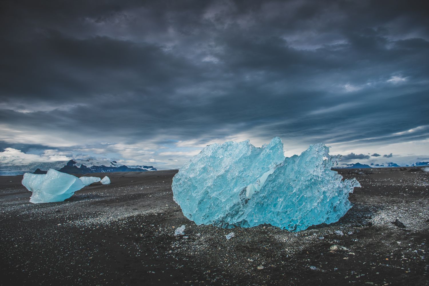 Striking blue glacial ice fragments rest on black sand beach under dramatic stormy skies in Iceland.