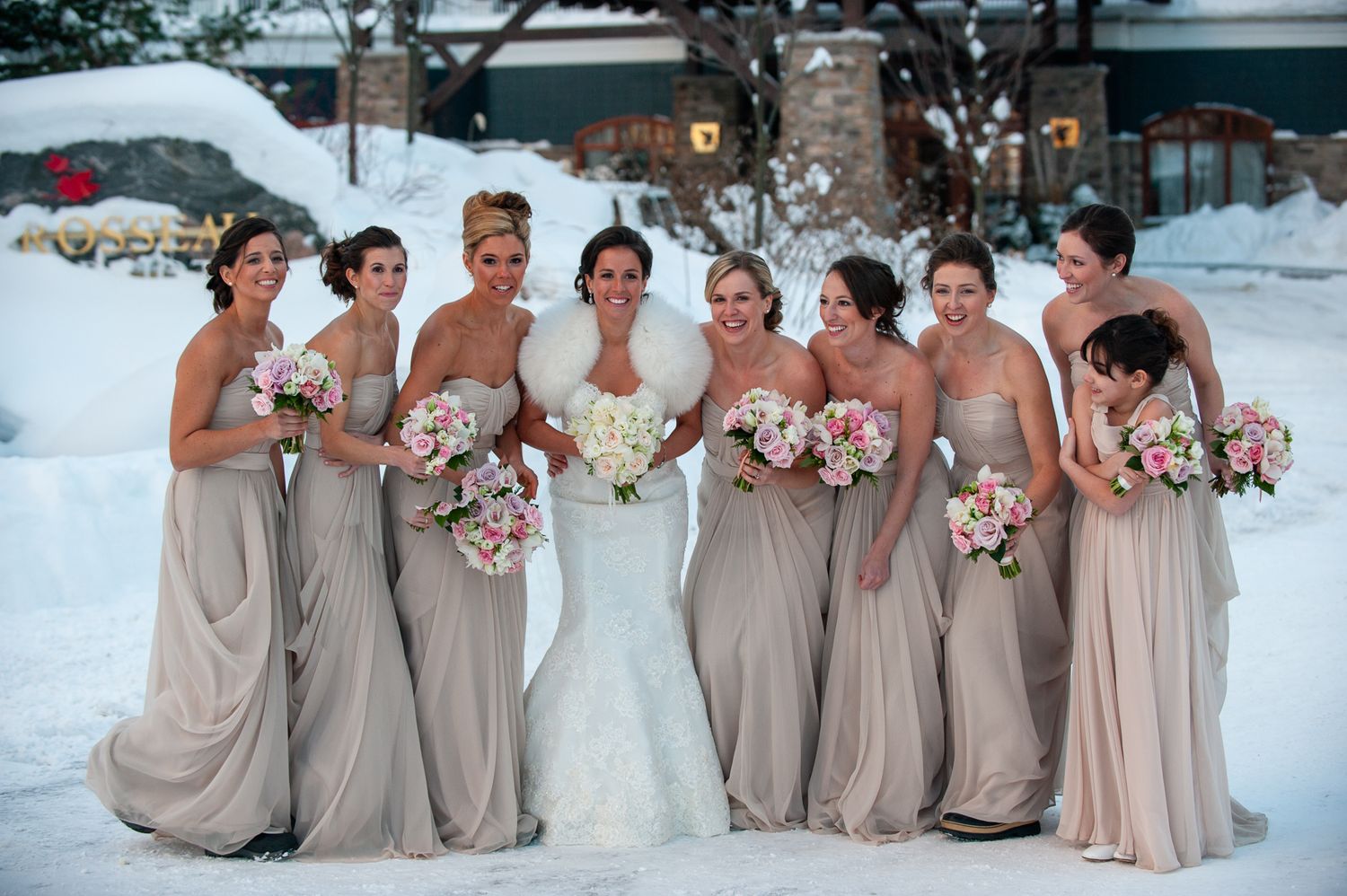 Bridal party wearing blush-colored gowns poses together in a snowy winter setting with pink bouquets.