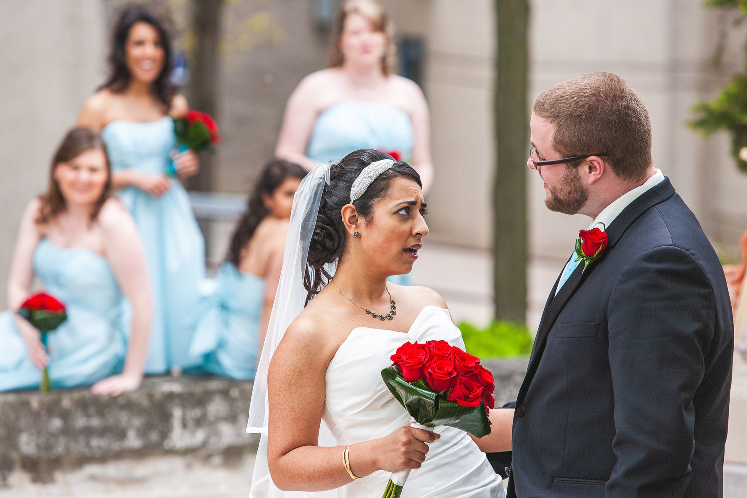 A bride in a white gown holds red roses while facing a groom in a dark suit as bridesmaids in blue dresses look on.