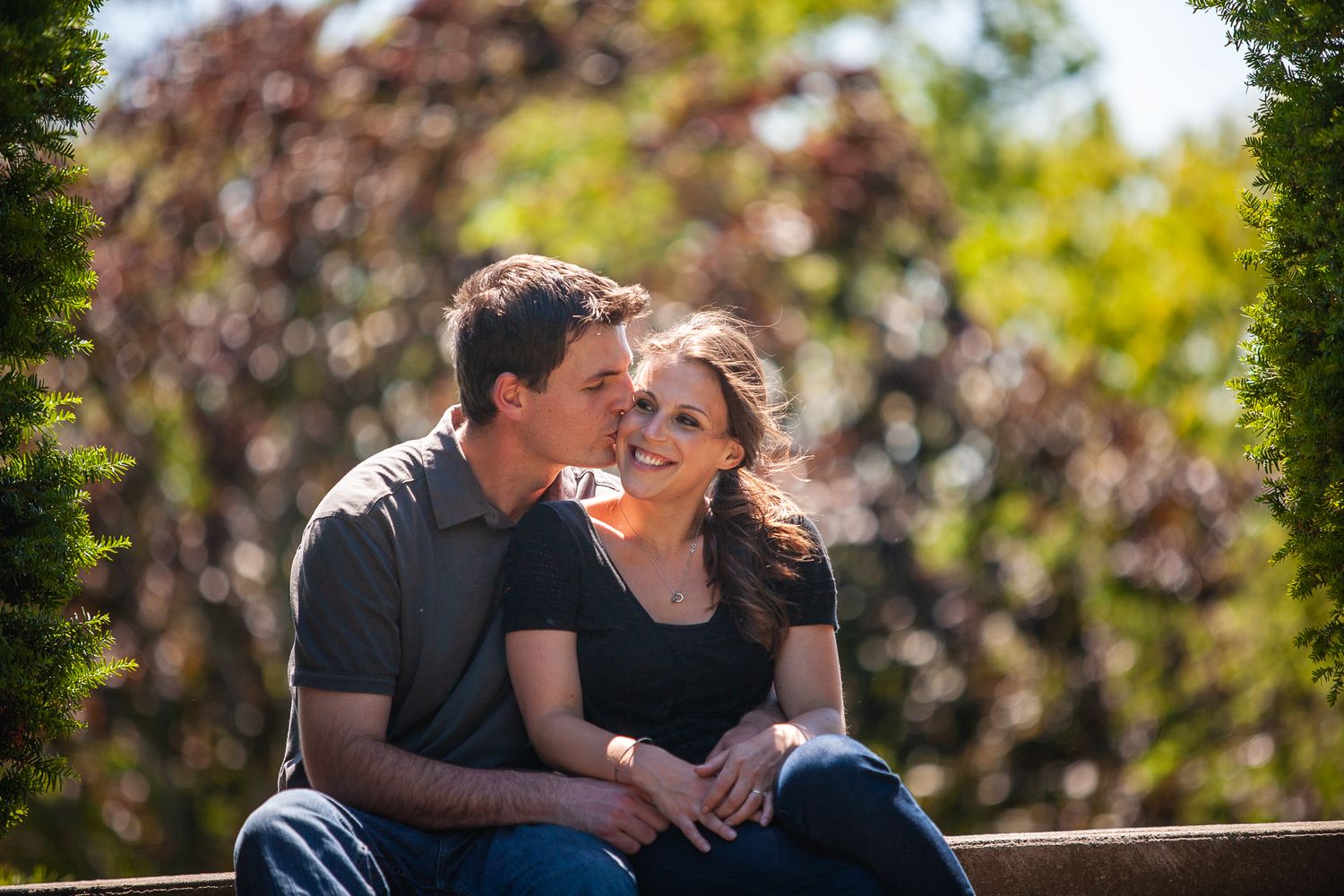 A romantic couple sits together on a bench in a garden setting during a sunny afternoon.