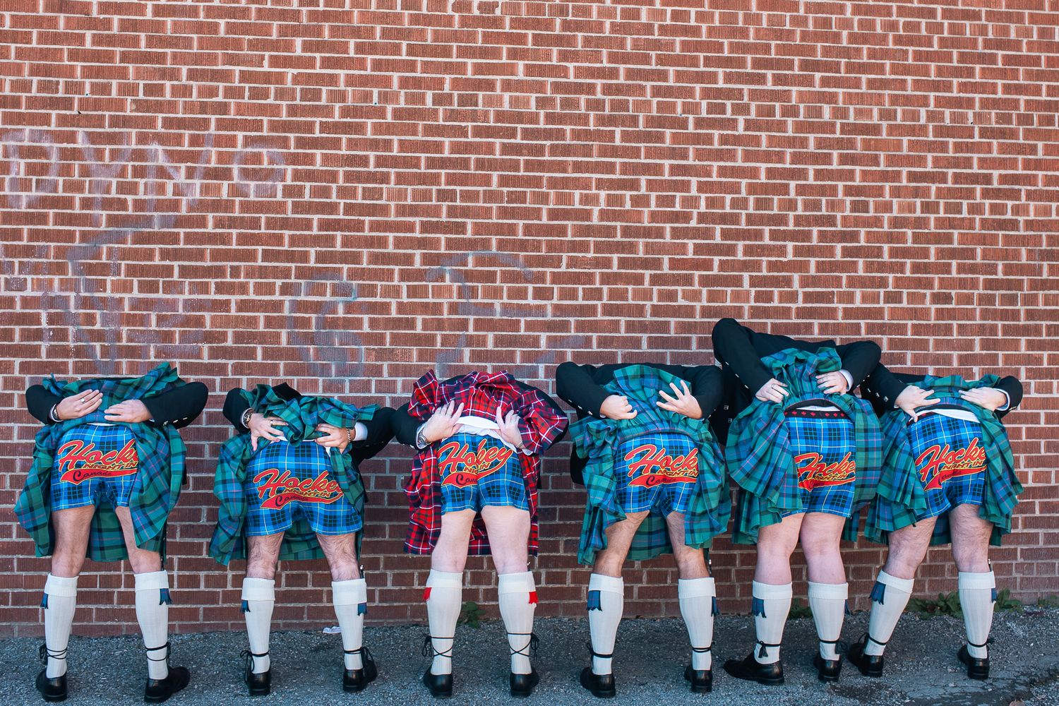 A lineup of kilted Scottish dancers leaning forward against a brick wall showing their colorful tartan uniforms from behind.