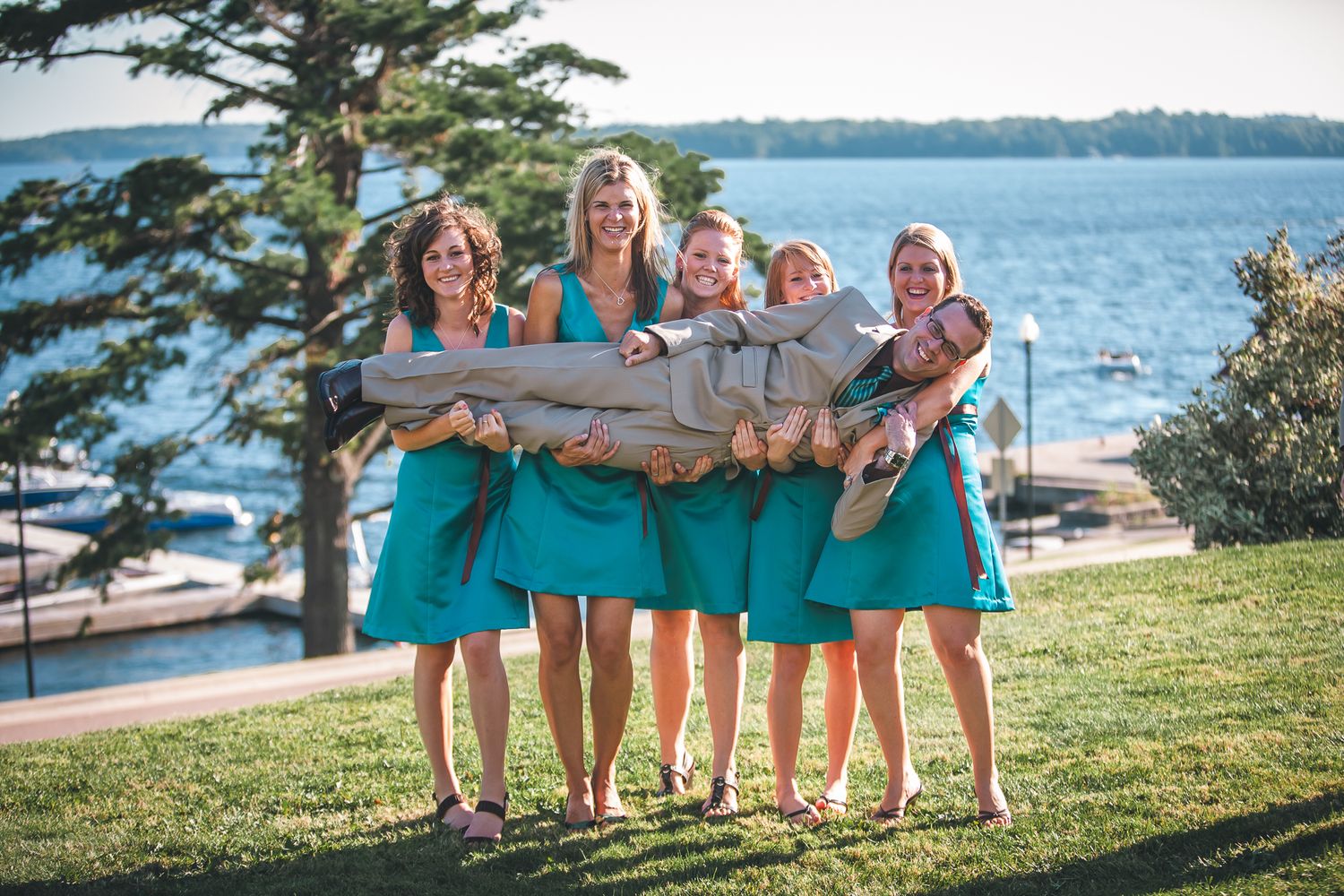 A group of bridesmaids in turquoise dresses laugh together on a grassy lawn overlooking a waterfront scene.