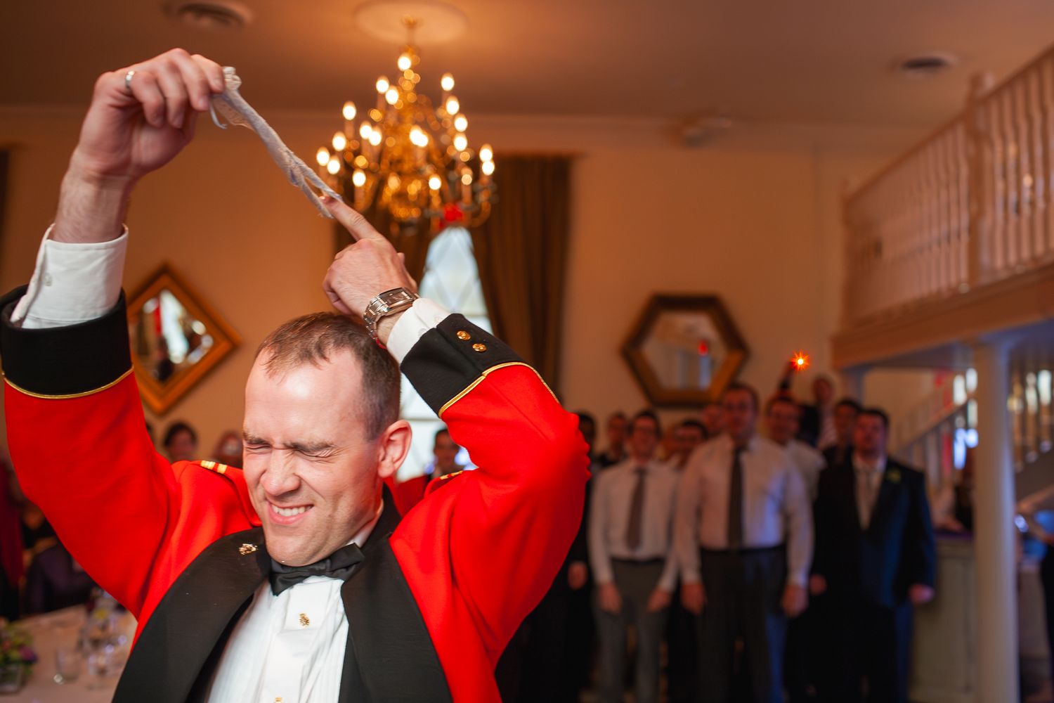 Man in tuxedo dancing joyfully at a wedding reception with guests and a chandelier in the background.