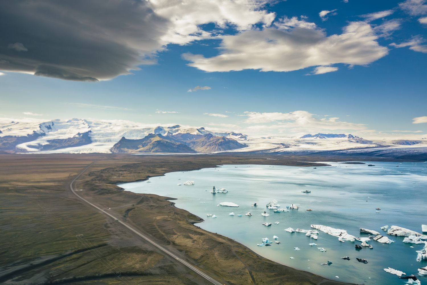 Glacier lagoon Jokulsarlon in Iceland with floating icebergs and winding road along shore under dramatic clouds.