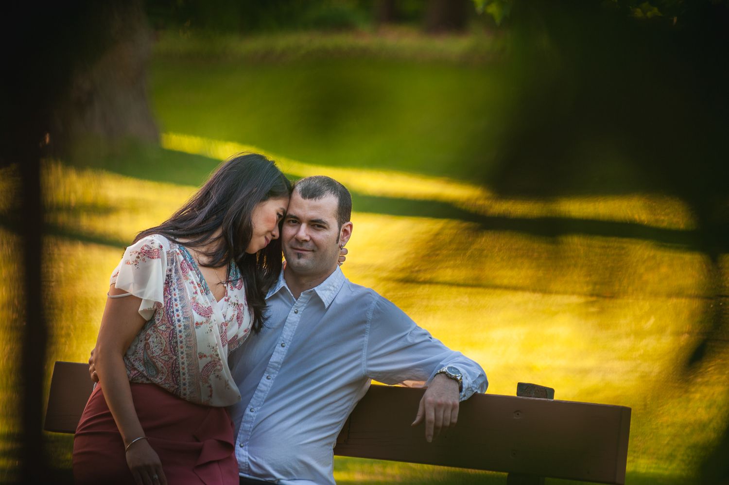 A romantic couple sits close together on a bench in a sunlit park with golden bokeh lighting in the background.
