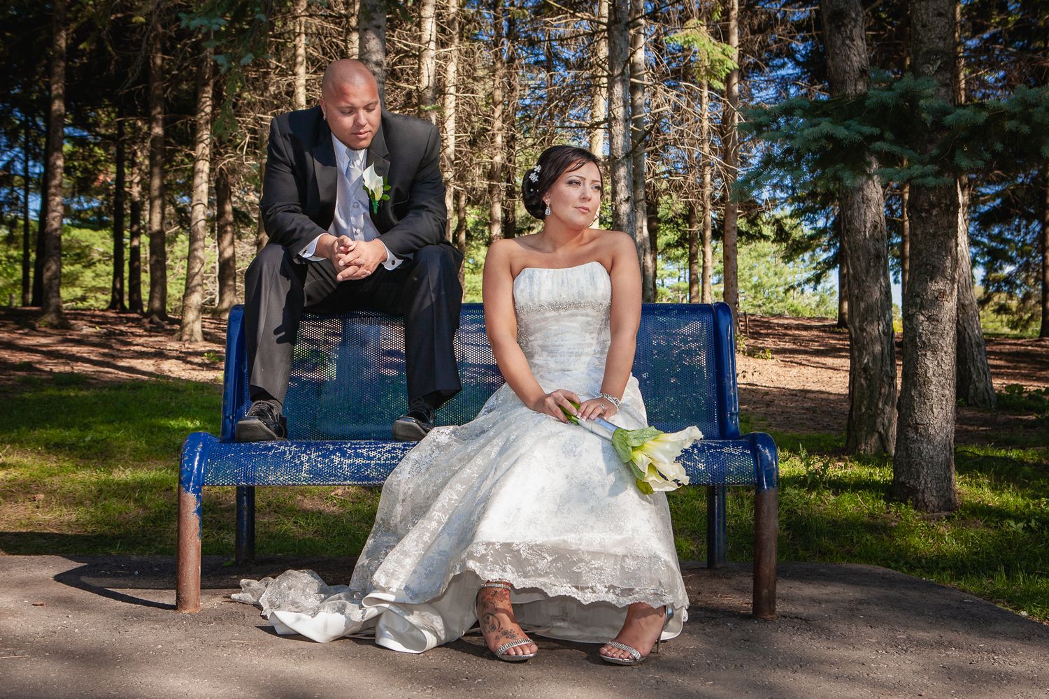 A couple in wedding attire sits on a blue bench surrounded by pine trees in an outdoor setting.