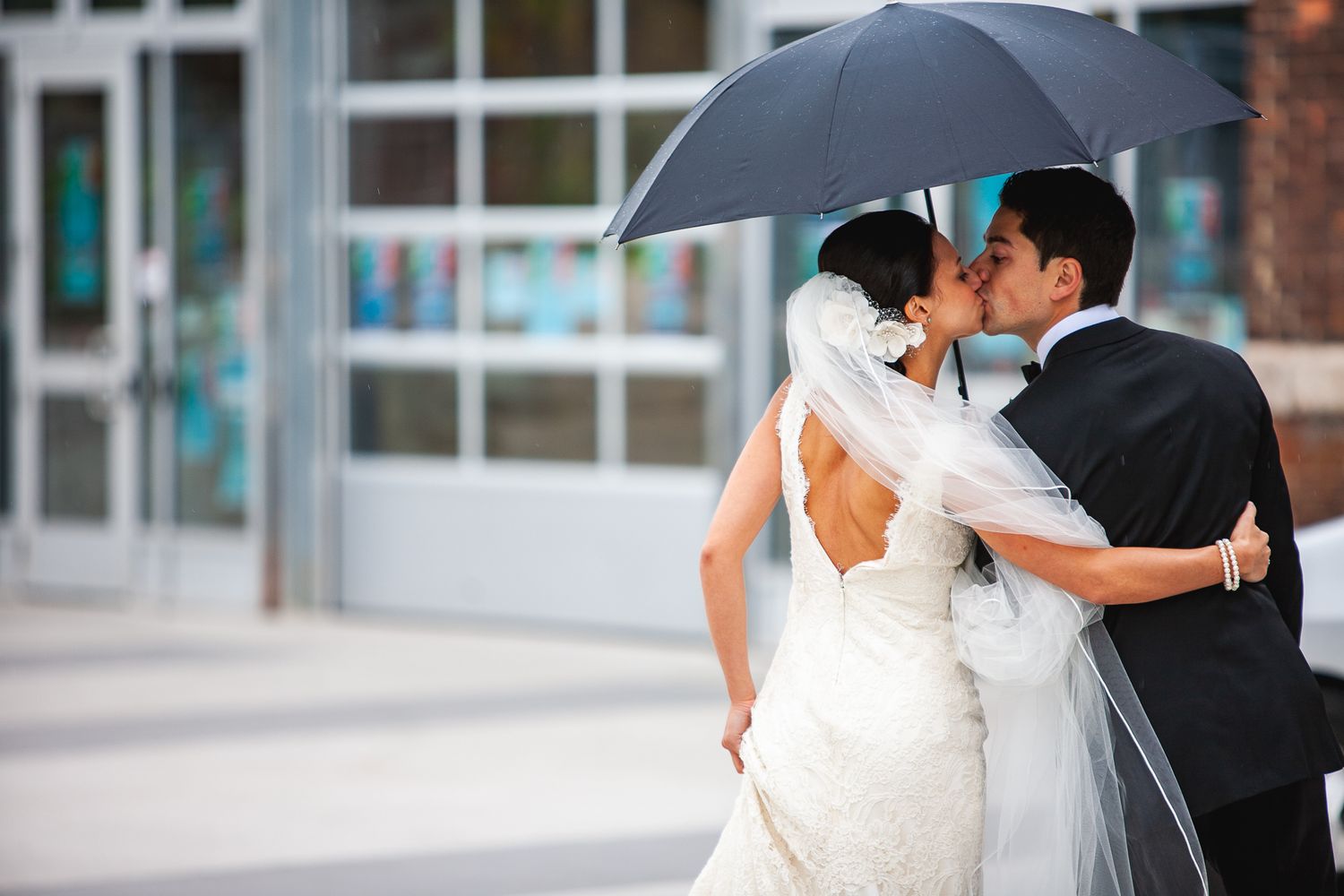 Romantic wedding couple shares an intimate moment under a black umbrella on a rainy day.