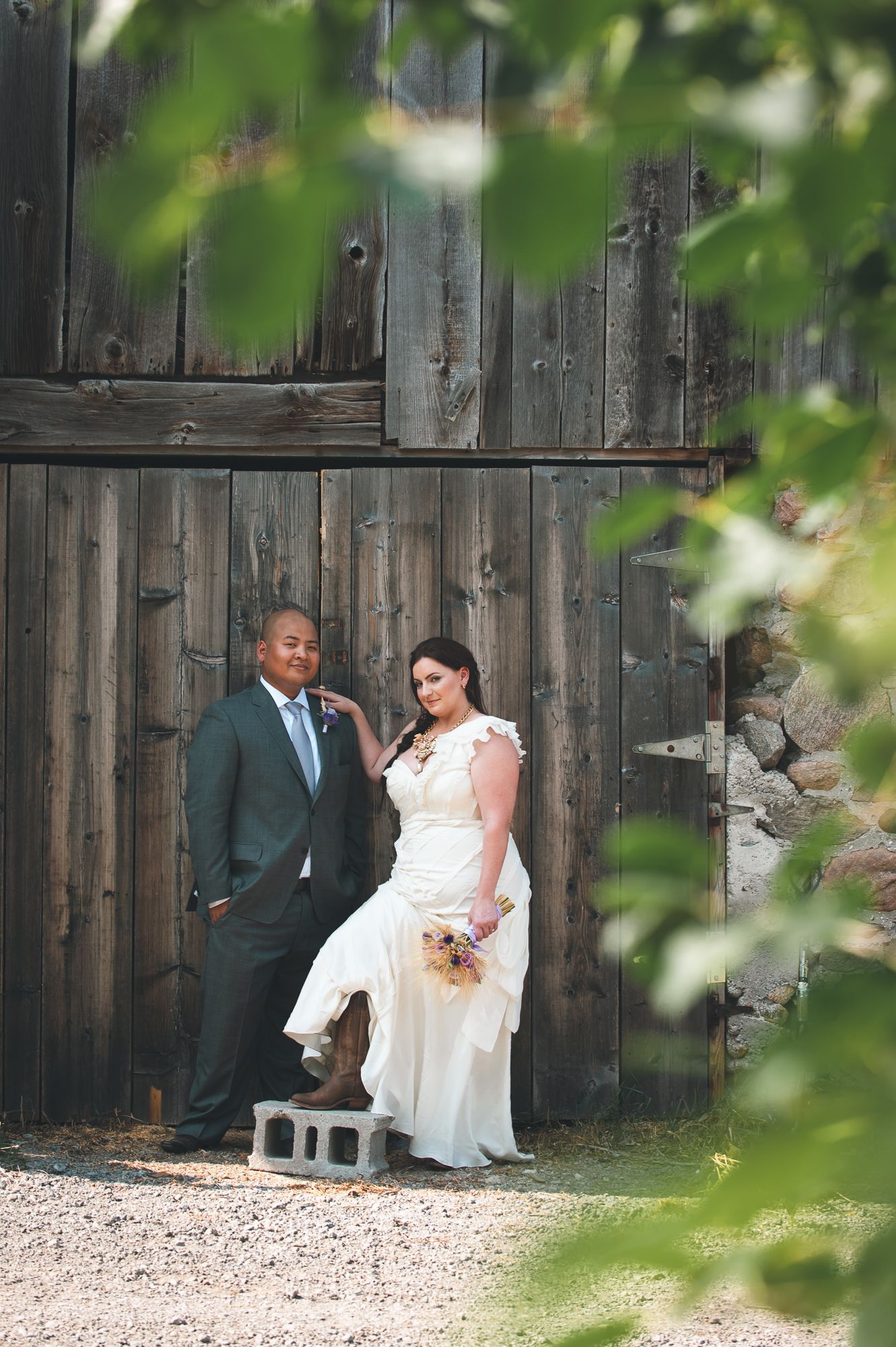 A rustic wedding couple poses against a weathered wooden barn wall framed by soft green leaves.