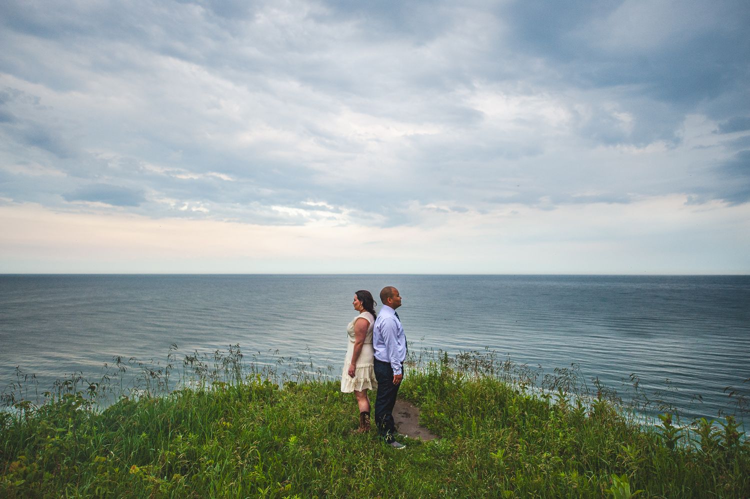 Two people stand on a grassy cliff overlooking the ocean under a cloudy summer sky.
