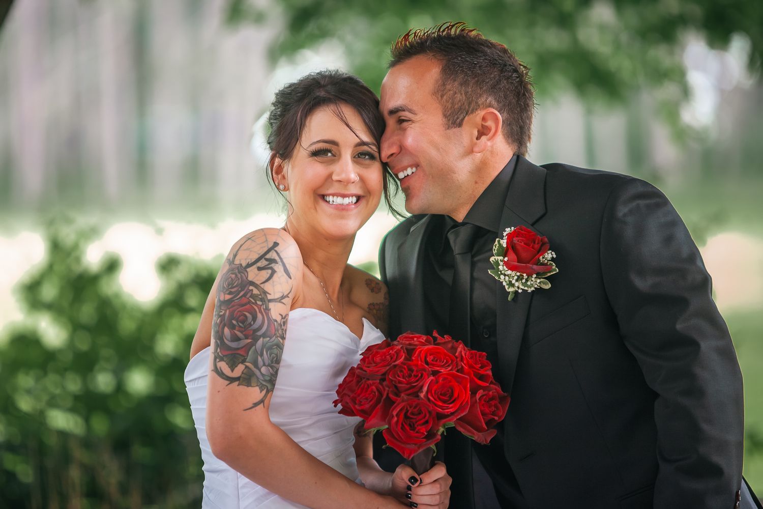 Happy couple shares intimate moment at outdoor wedding with red rose bouquet and matching boutonniere.