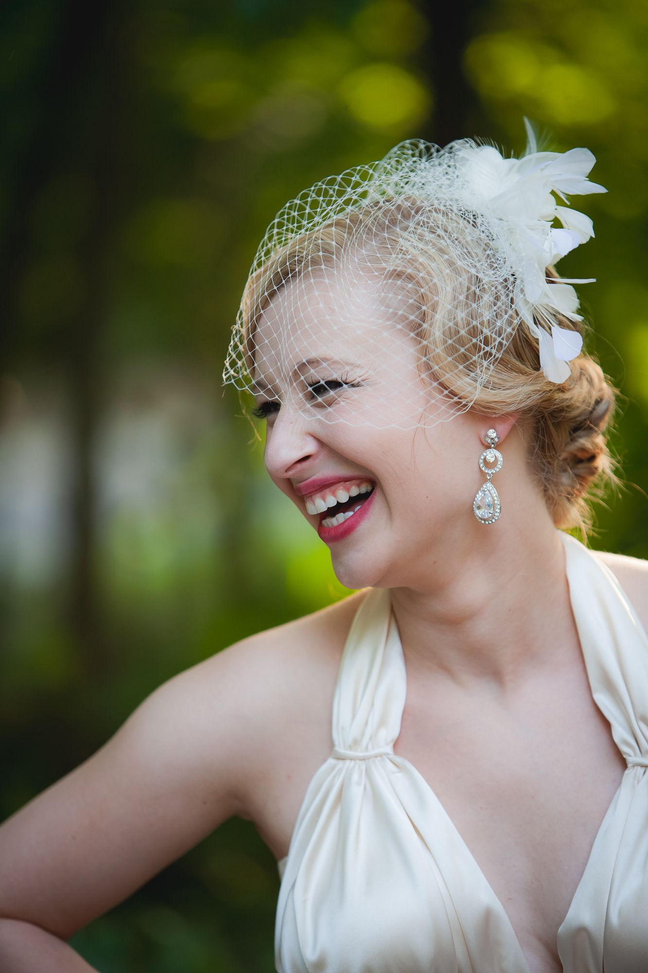 A joyful bride wearing a classic white halter dress, birdcage veil, and crystal drop earrings laughs during outdoor photos.