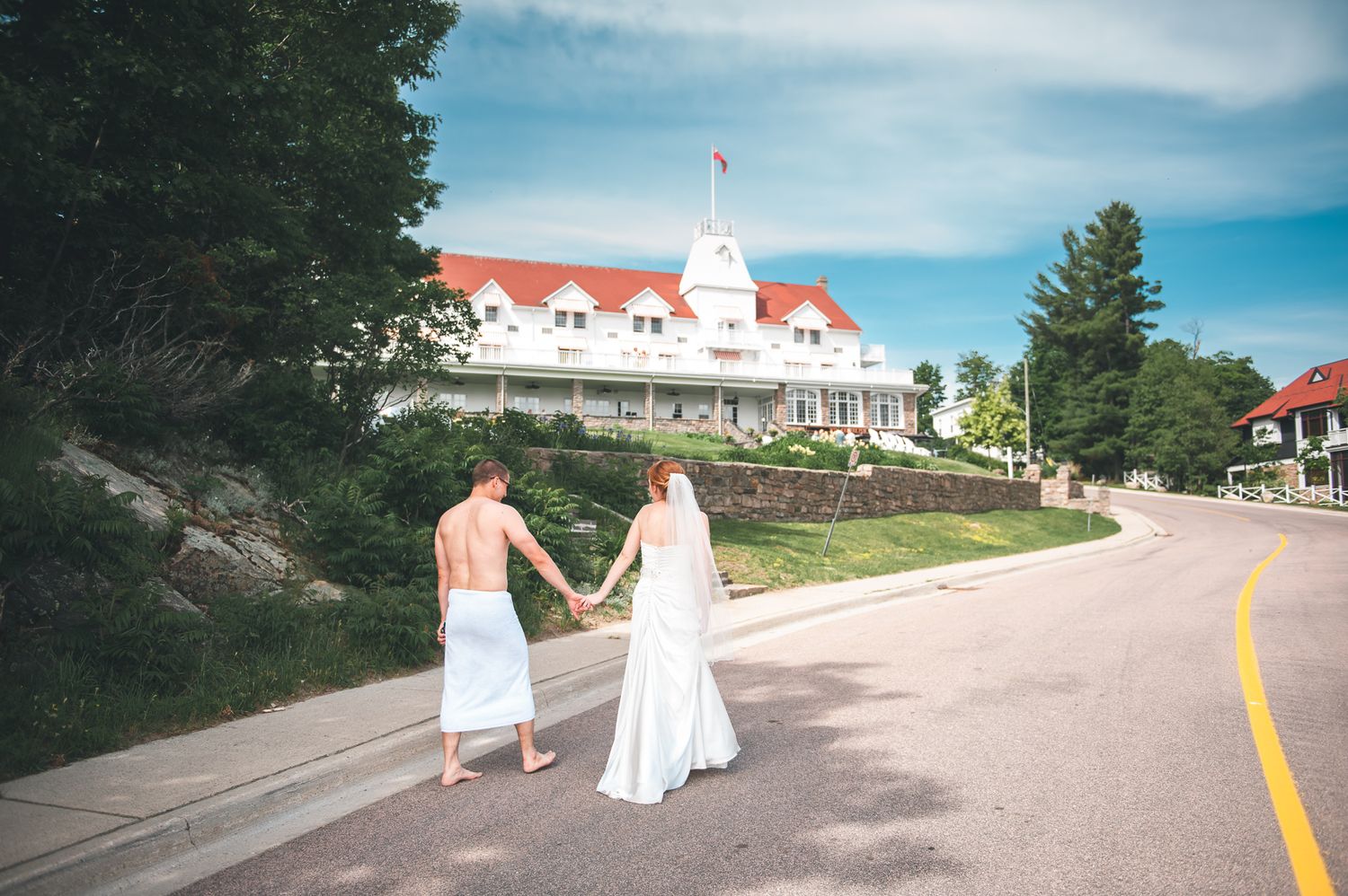 Two people holding hands walk down a road towards a grand white hotel with red roofs on a sunny summer day.