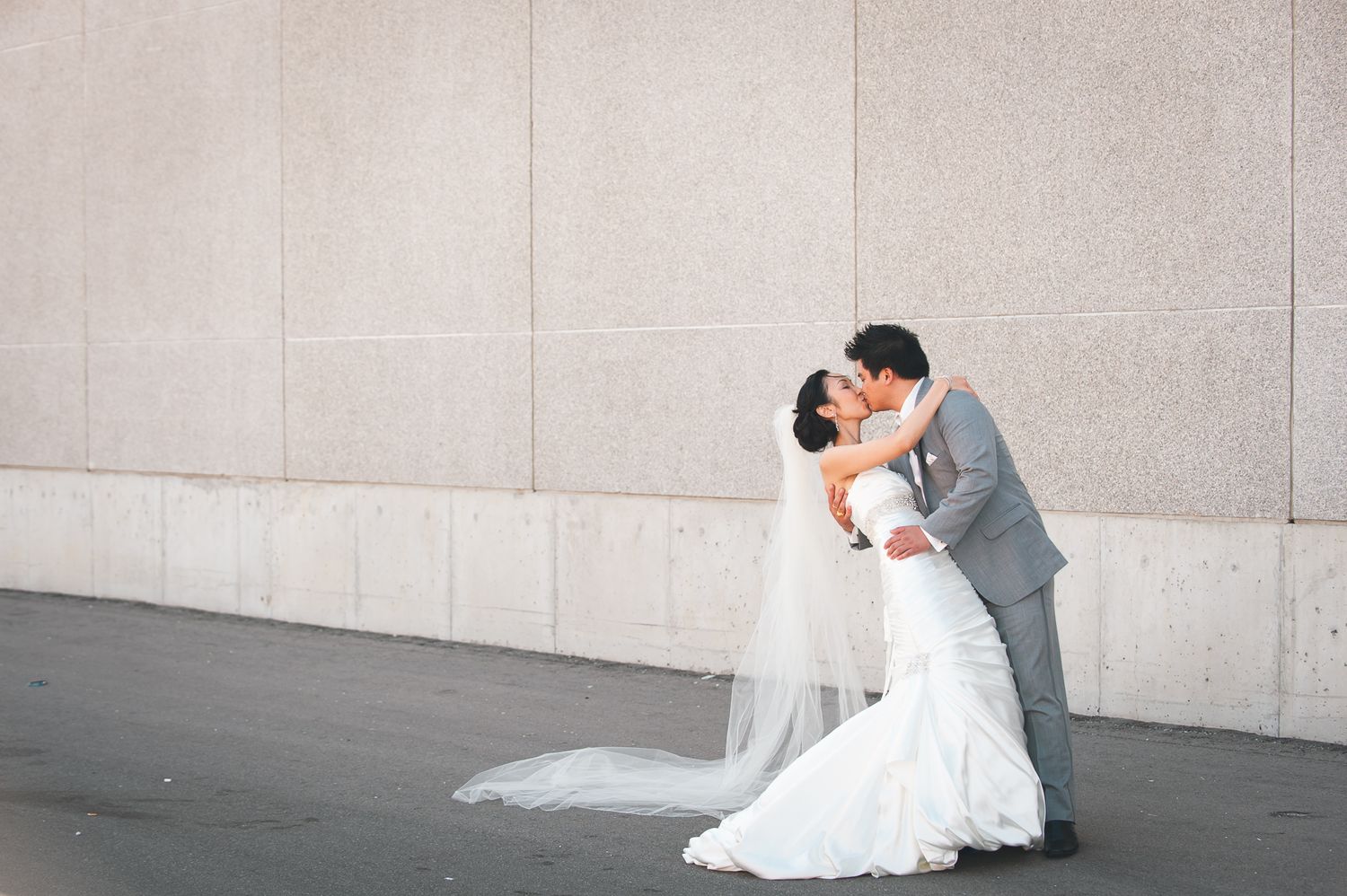 A couple in wedding attire embrace and kiss against a minimalist gray wall backdrop during their wedding photoshoot.