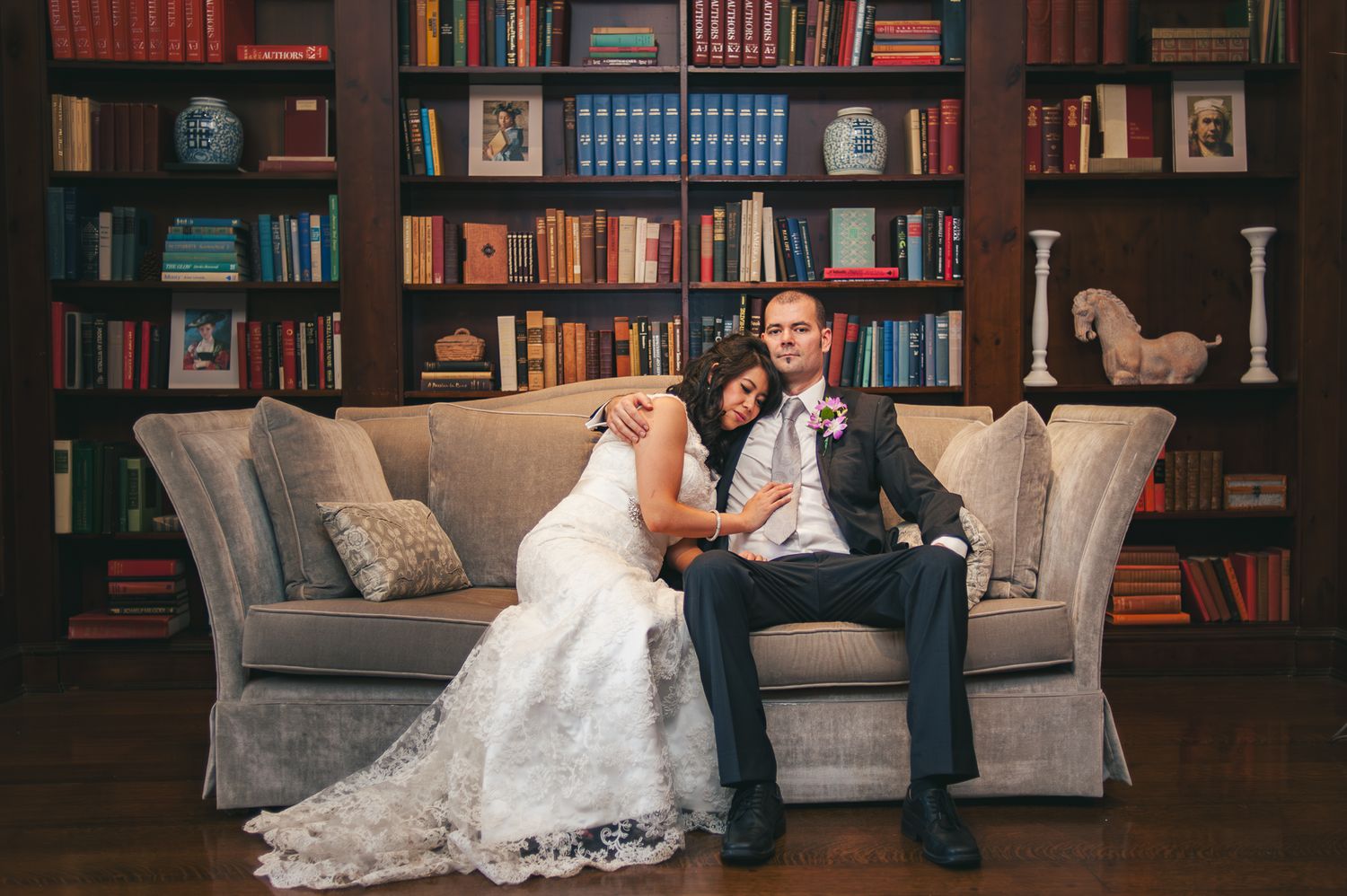 Couple in wedding attire sits together on gray couch in library with floor-to-ceiling bookshelves.