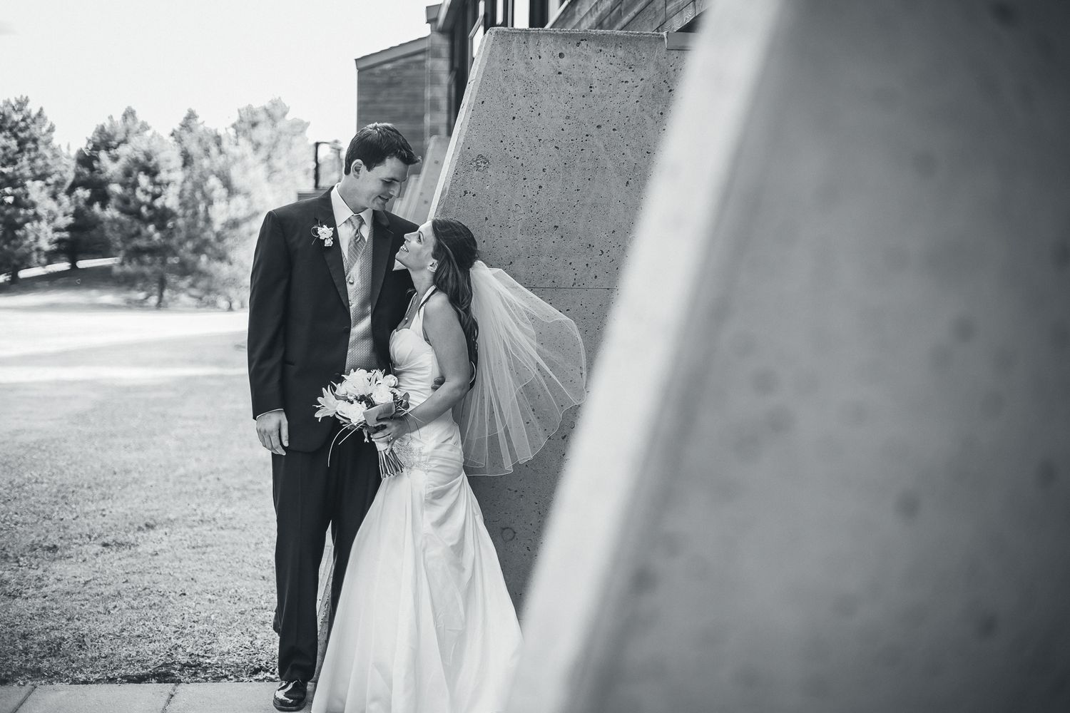 Black and white wedding photo of a couple in formal attire standing against a wall with an umbrella parasol.