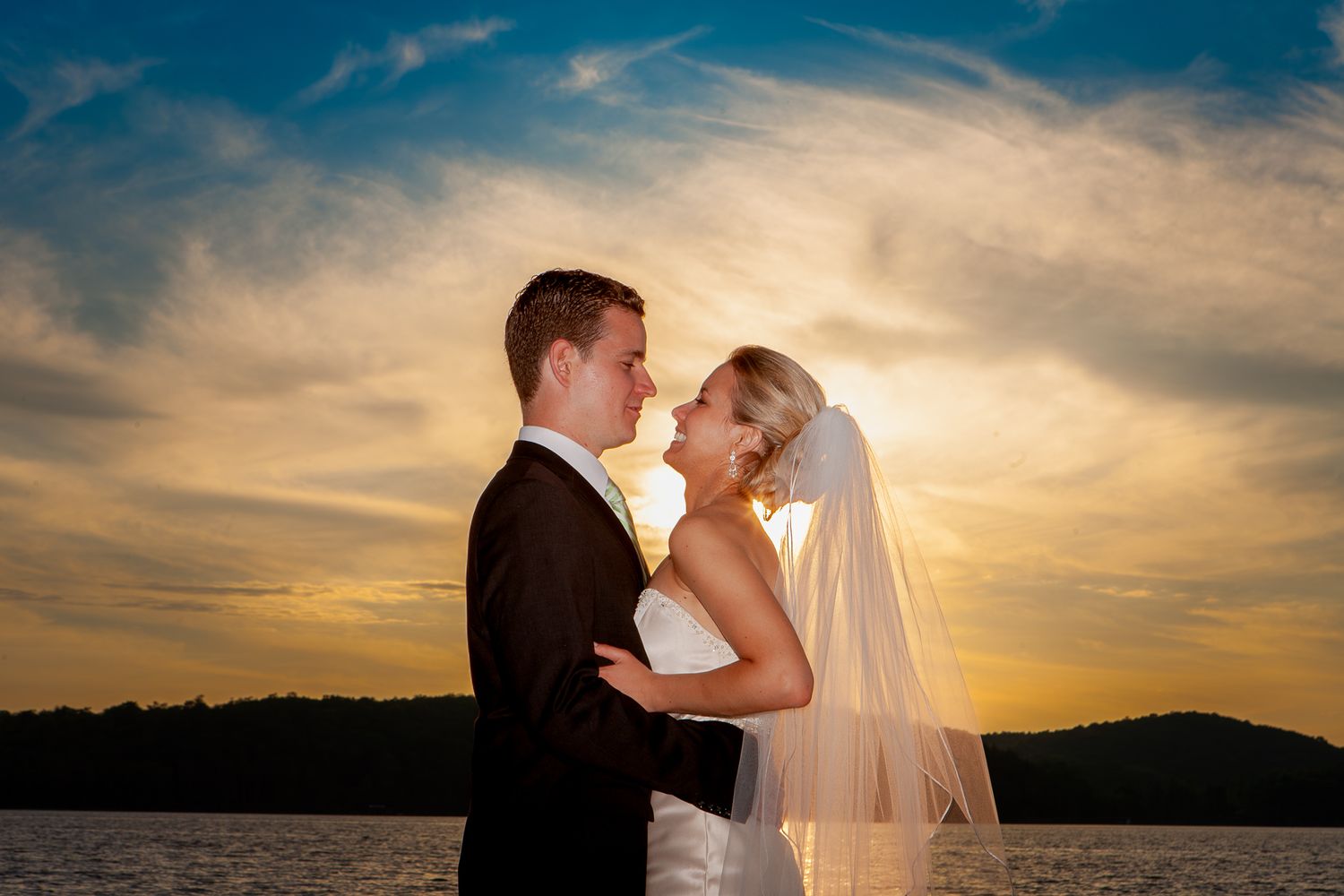 A romantic sunset wedding photo silhouetted against golden clouds and mountains by a lake.