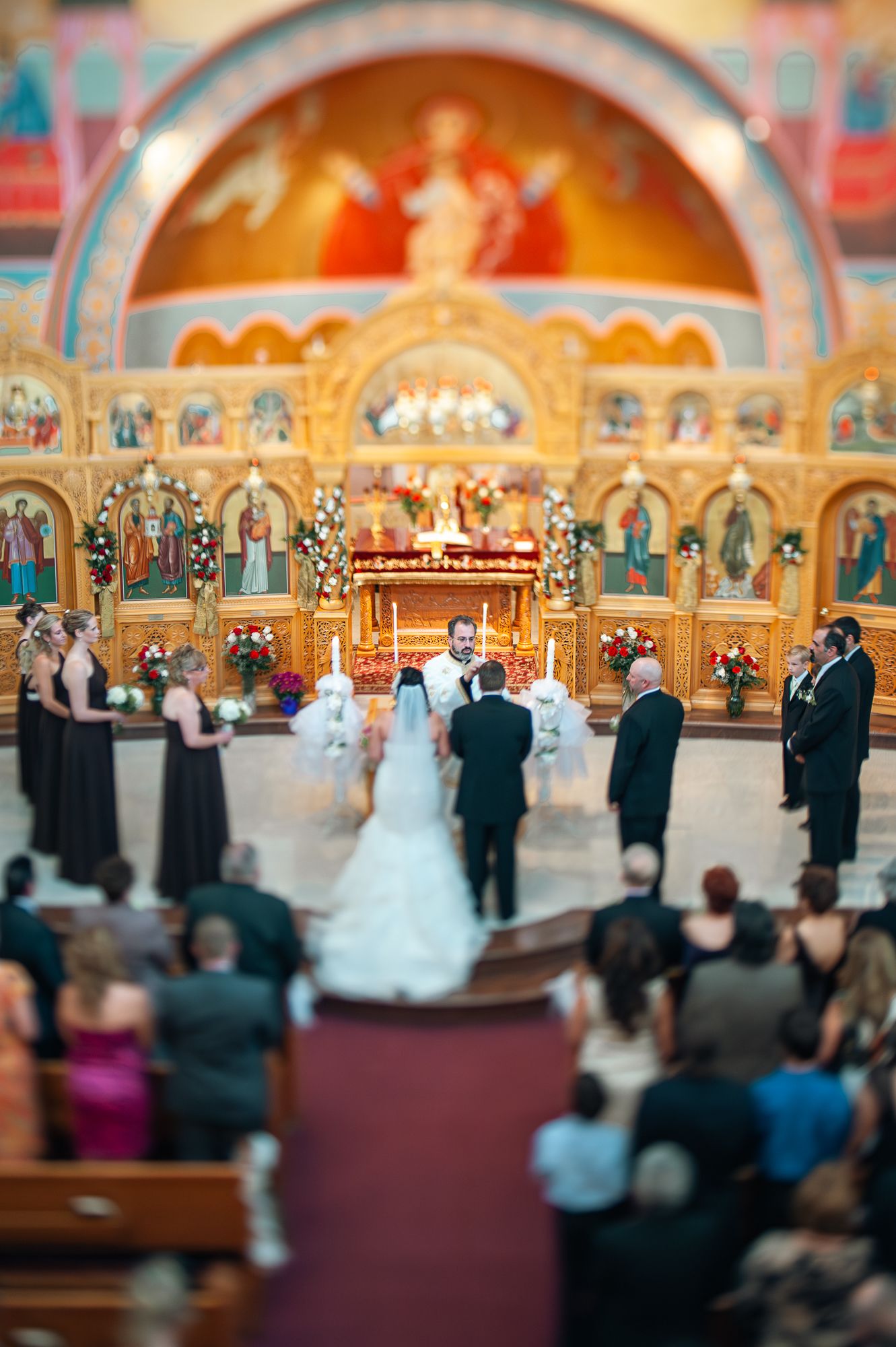 Orthodox Christian wedding ceremony taking place in ornate church with gold iconostasis and red carpeted aisle.