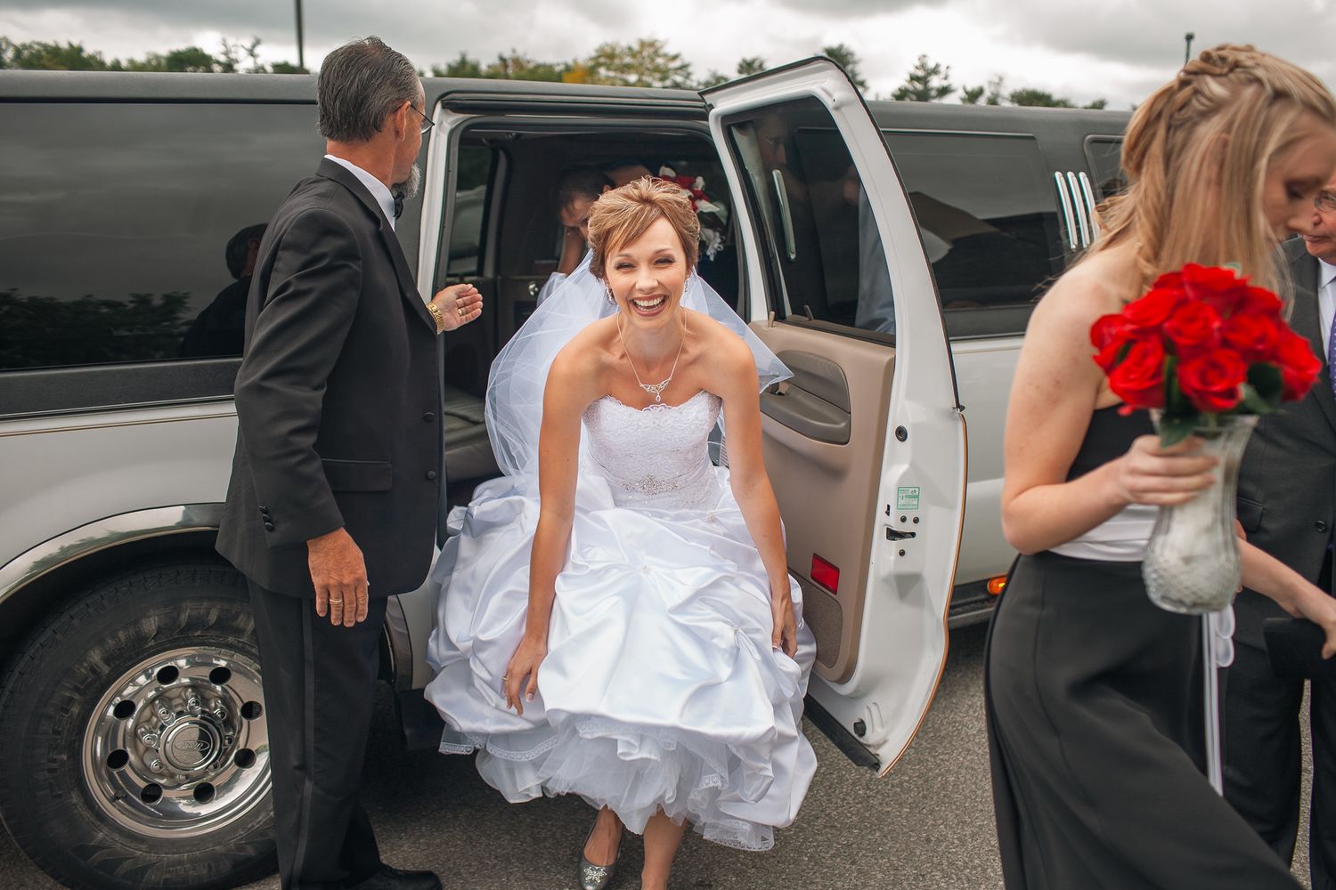 Someone in a white wedding dress steps out of a limousine while others in formal attire assist nearby.