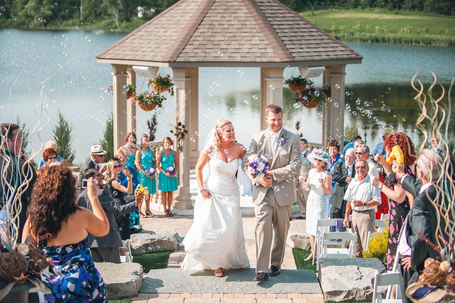Outdoor wedding ceremony taking place at a gazebo overlooking a serene lake on a sunny day.