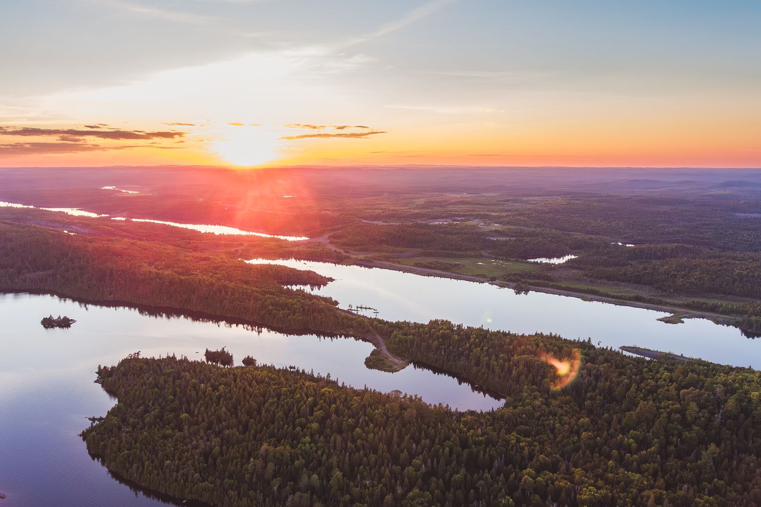 Aerial view of sunset over a winding lake surrounded by dense forest landscape with golden light reflecting on calm waters.