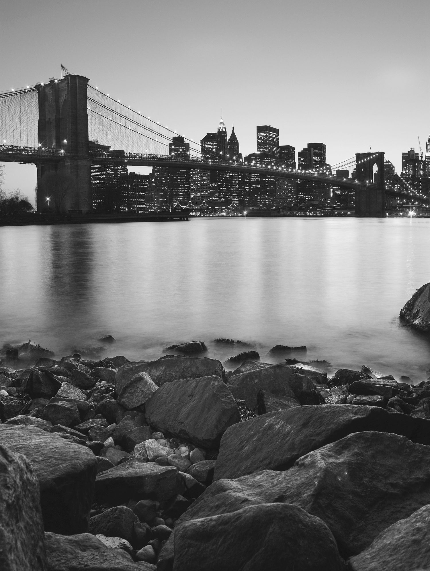 Black and white cityscape of Brooklyn Bridge and Manhattan skyline with rocky shoreline in the foreground.