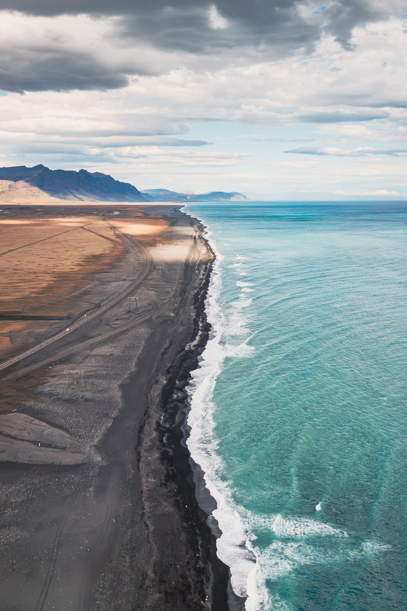 Dramatic aerial view of turquoise ocean waves meeting black sand beach along a coastal road.