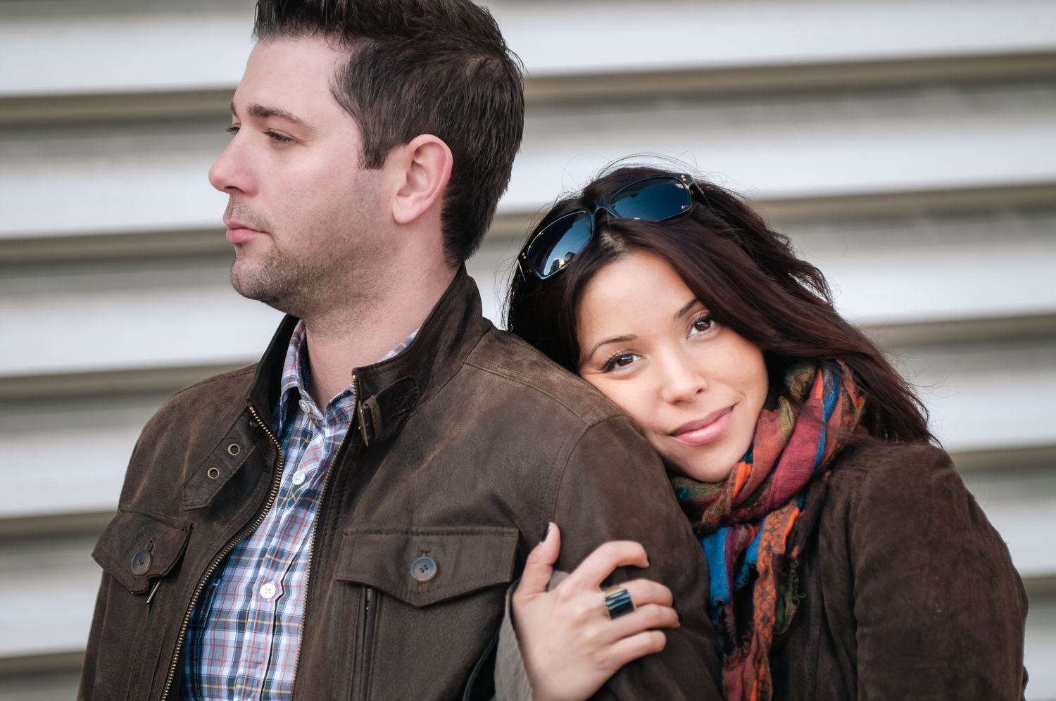 A couple in casual winter wear pose together against a white industrial wall in soft natural lighting.