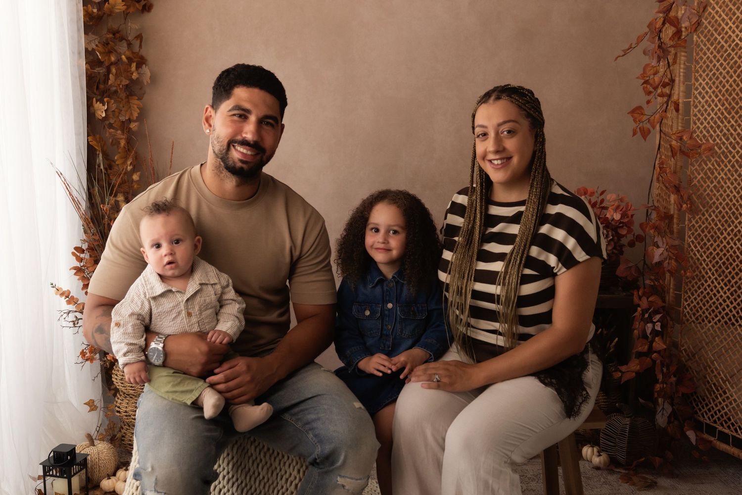 Family portrait with parents and two young children posed together in a warm autumn-themed studio setting.