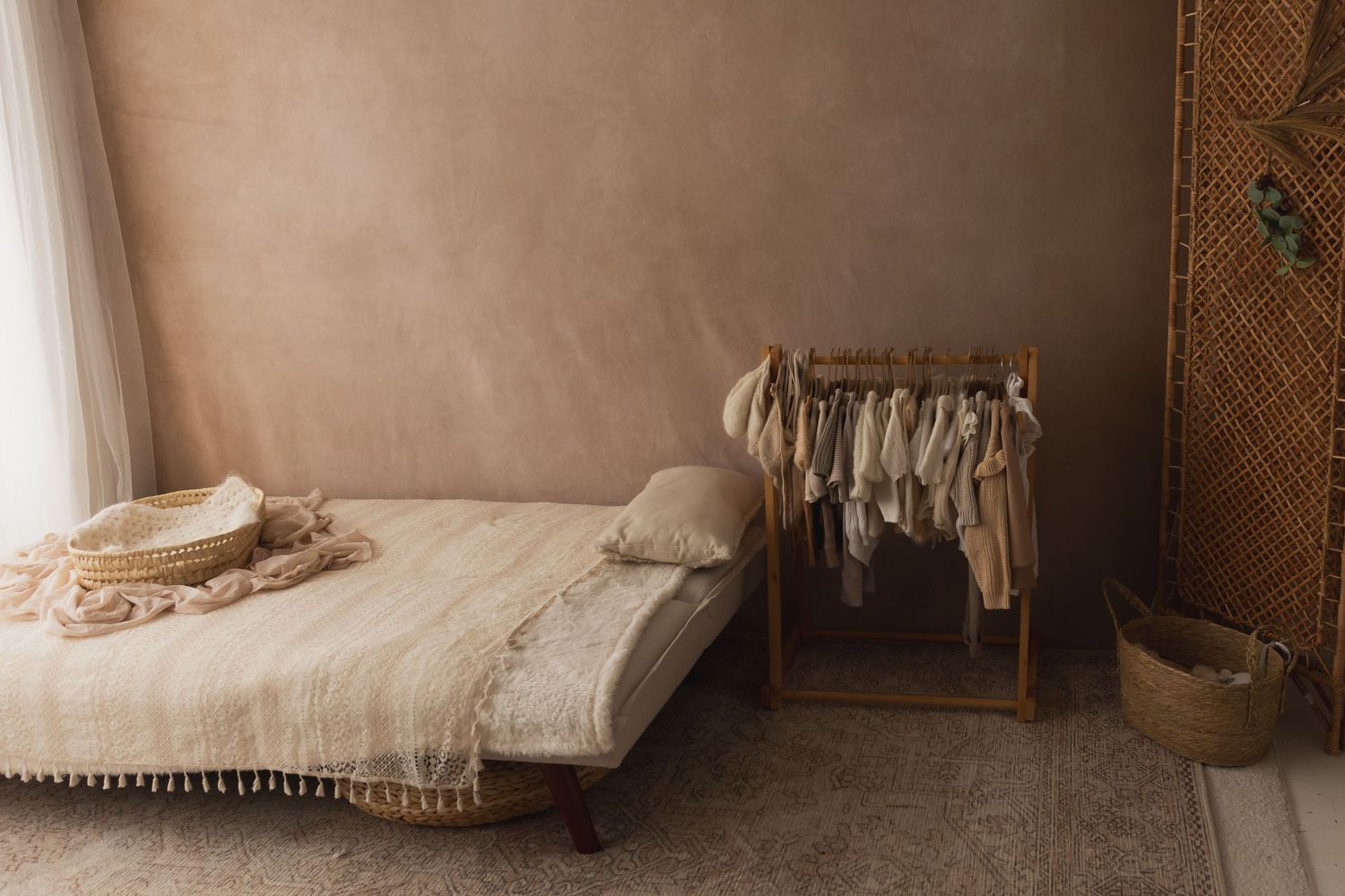 Boho nursery with a rattan clothes rack filled with neutral baby outfits, a low wooden bed with fringe blanket, and wicker basket.