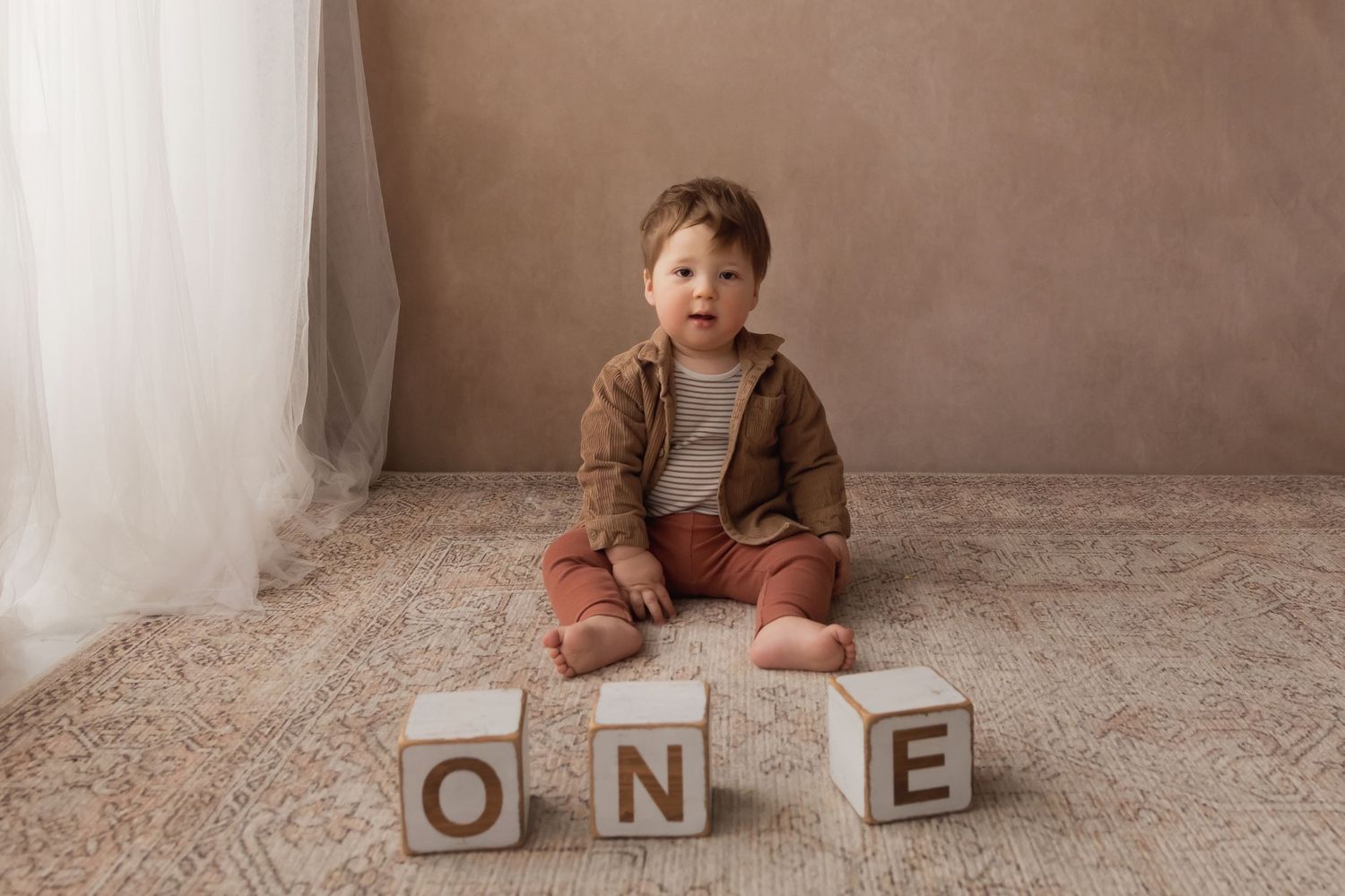 Happy baby boy in brown jacket and rust pants sits beside stacked ONE blocks for first birthday photos.