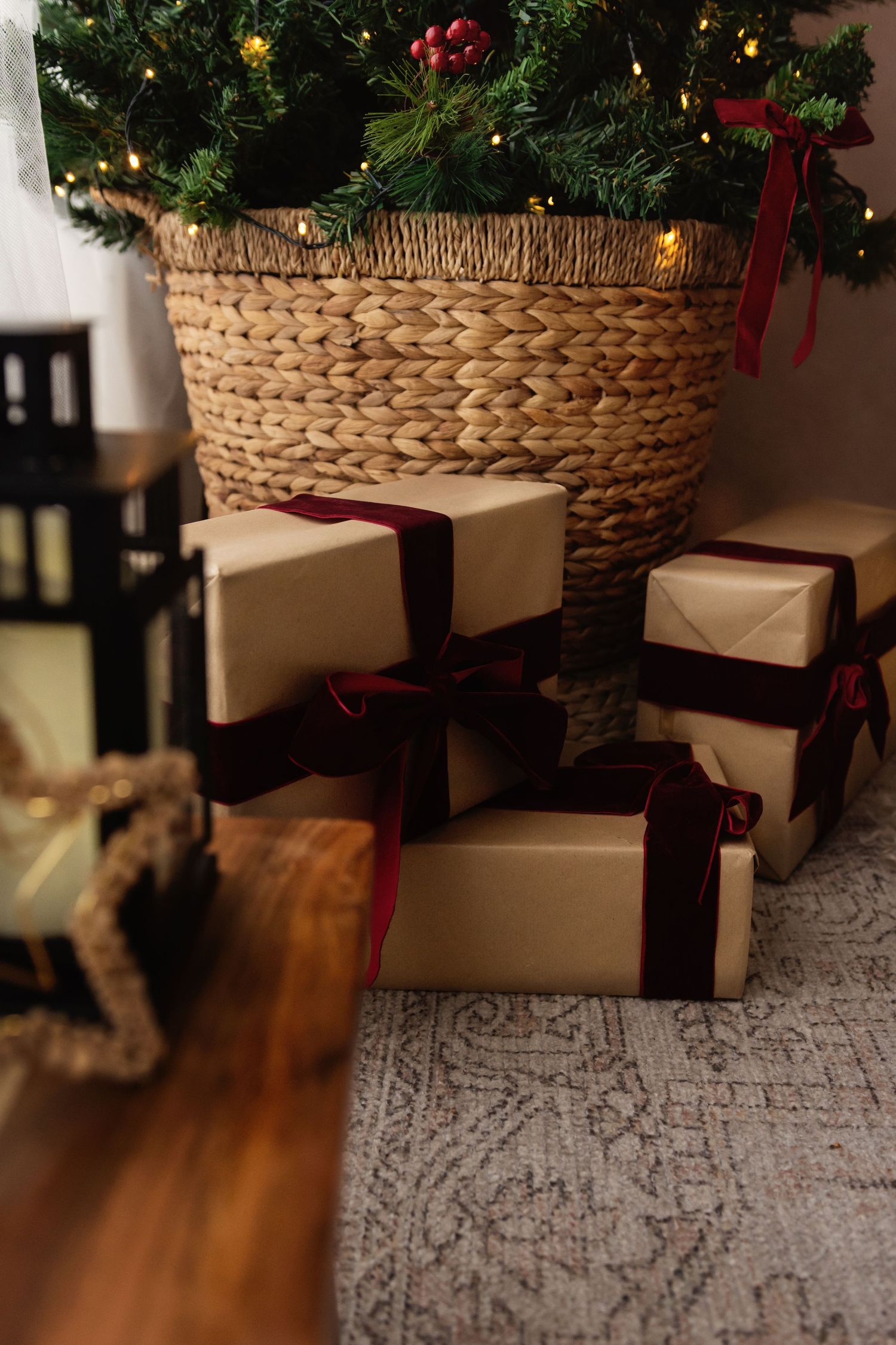 Christmas gifts wrapped in kraft paper with burgundy ribbons arranged under a decorated tree in a wicker basket.