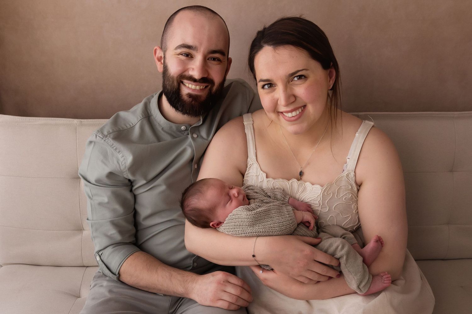 Smiling new parents sitting on couch together holding their swaddled newborn baby.