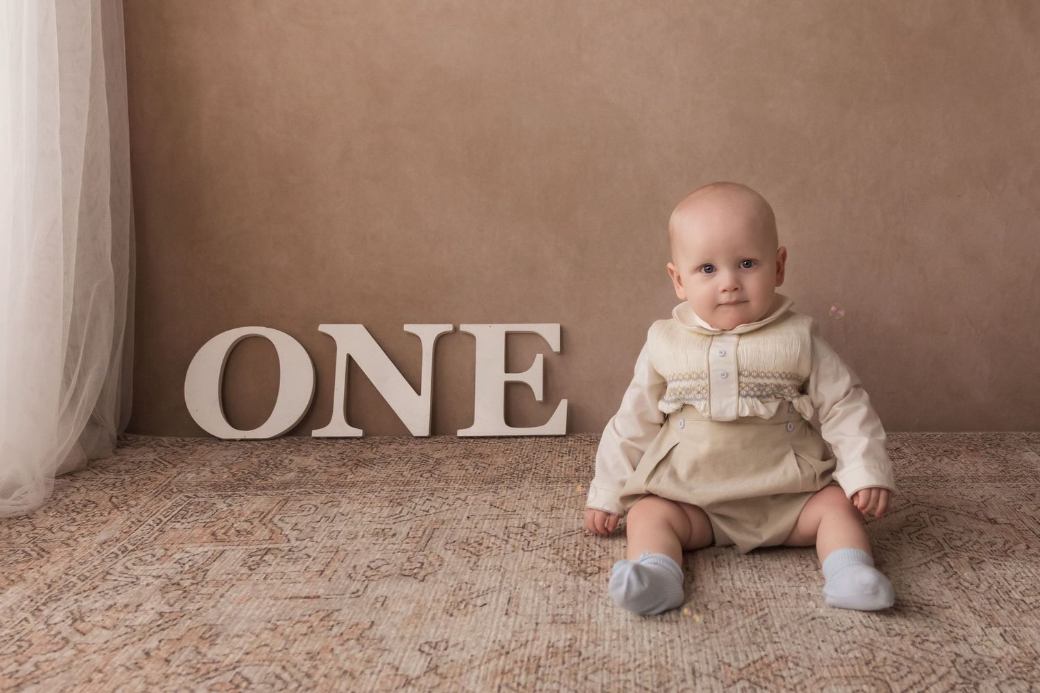 Adorable baby boy in cream outfit sits beside large white ONE letters for first birthday.