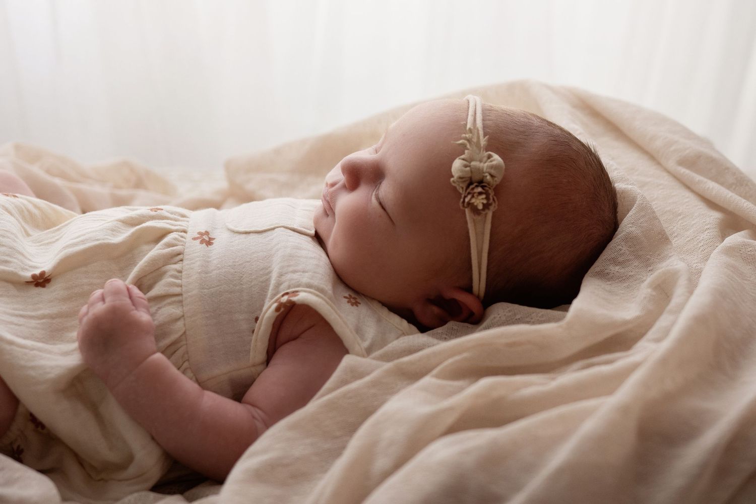 Peaceful newborn baby girl with floral headband sleeping on cream blanket.