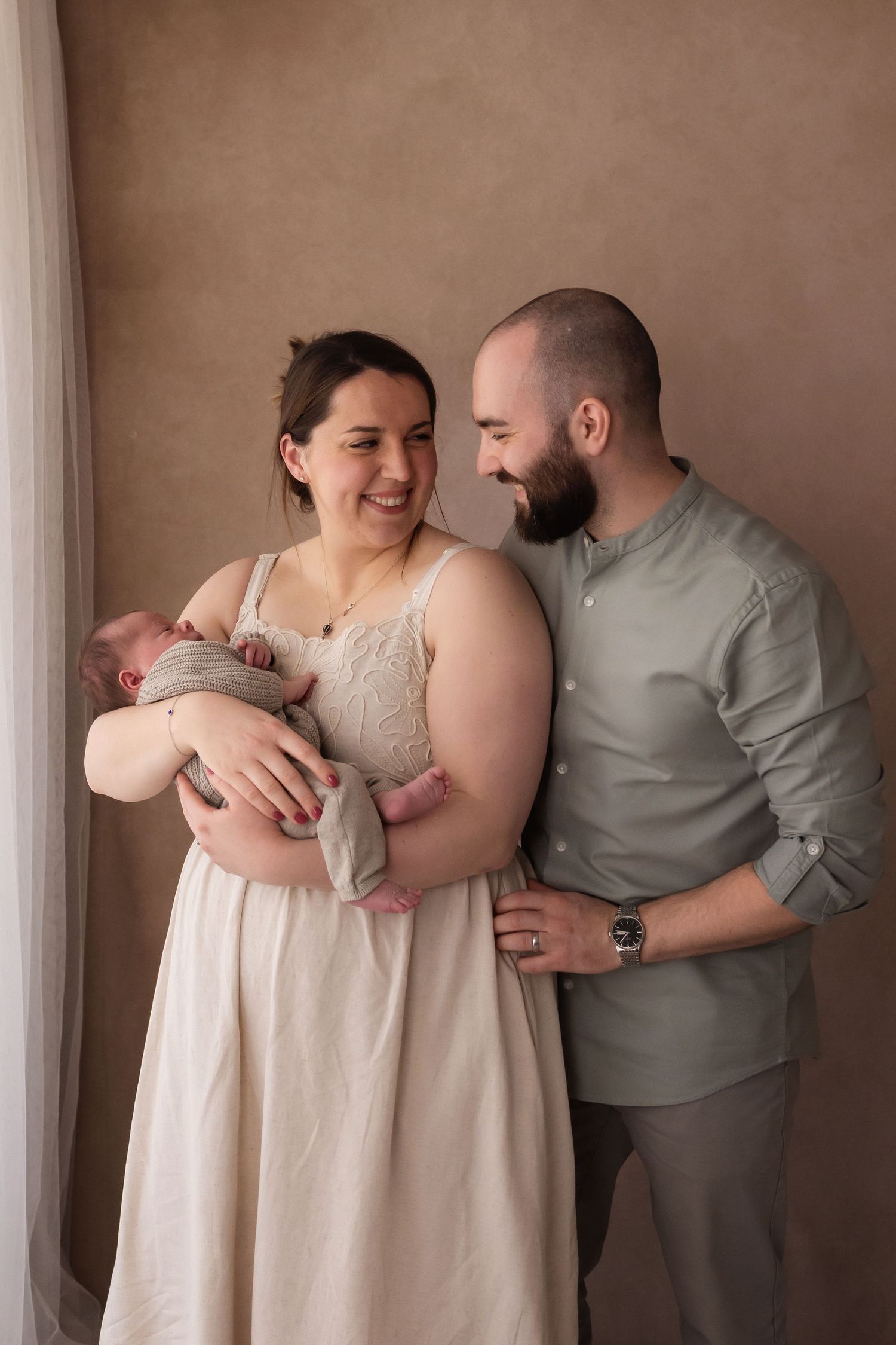 Happy new parents smiling at each other while mother holds newborn baby in studio.