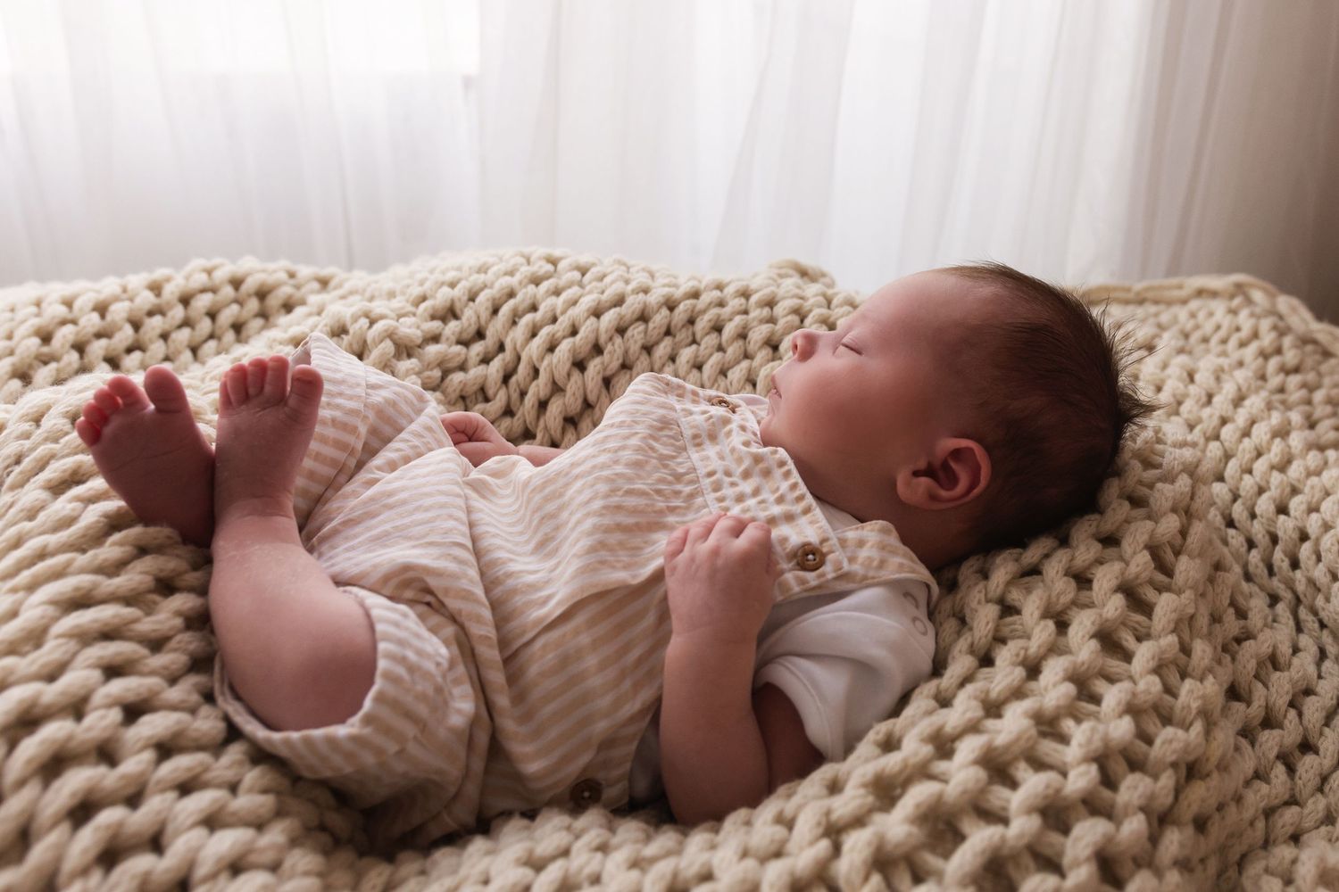 Peaceful newborn baby sleeping on a chunky cream knit blanket wearing a beige knit romper.