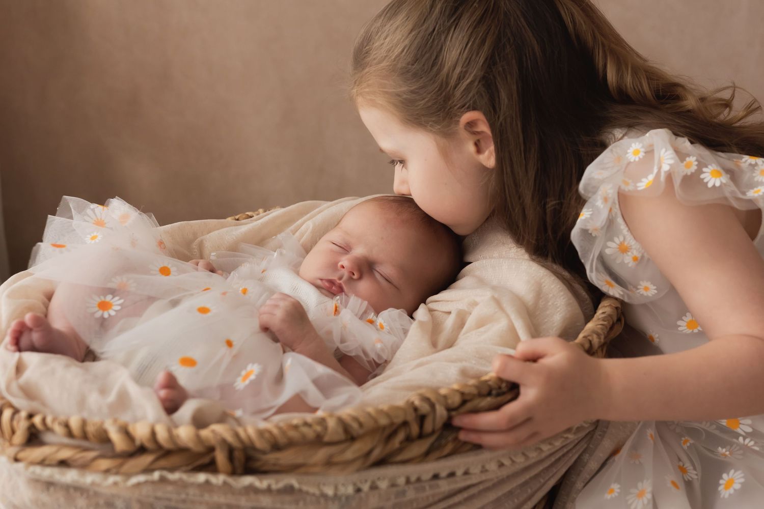 Young girl in daisy dress kissing sleeping newborn sibling in a wicker Moses basket.