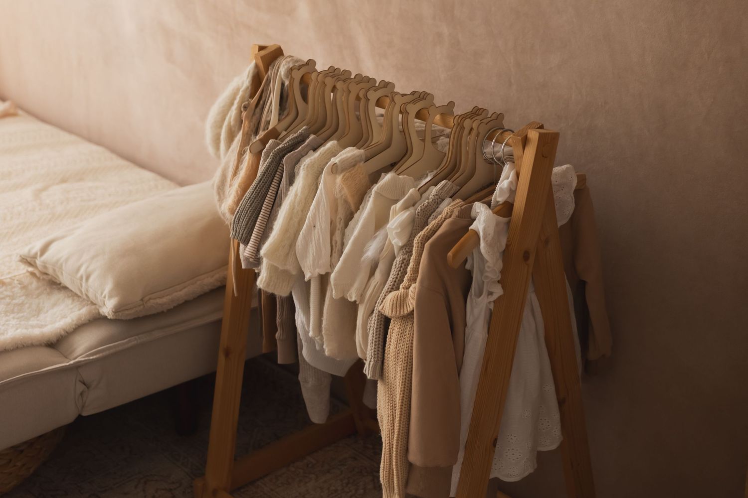Neutral-toned baby clothes hanging on a wooden clothing rack beside a cozy beige sofa.