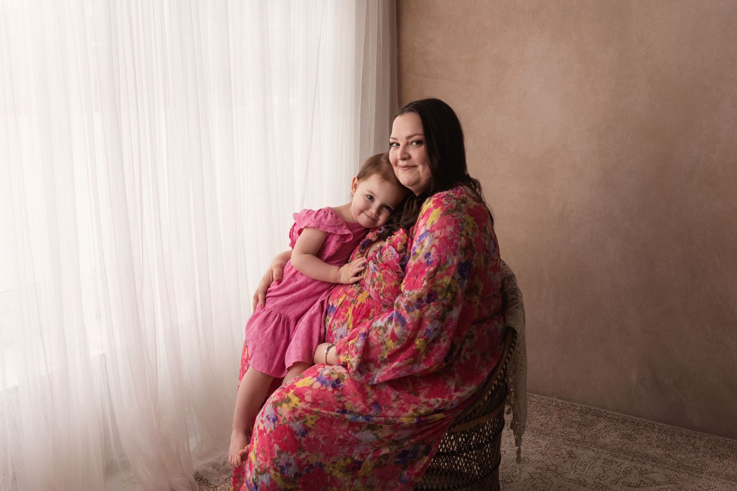 Pregnant woman in a pink floral dress sitting in a chair lovingly embracing her young daughter.