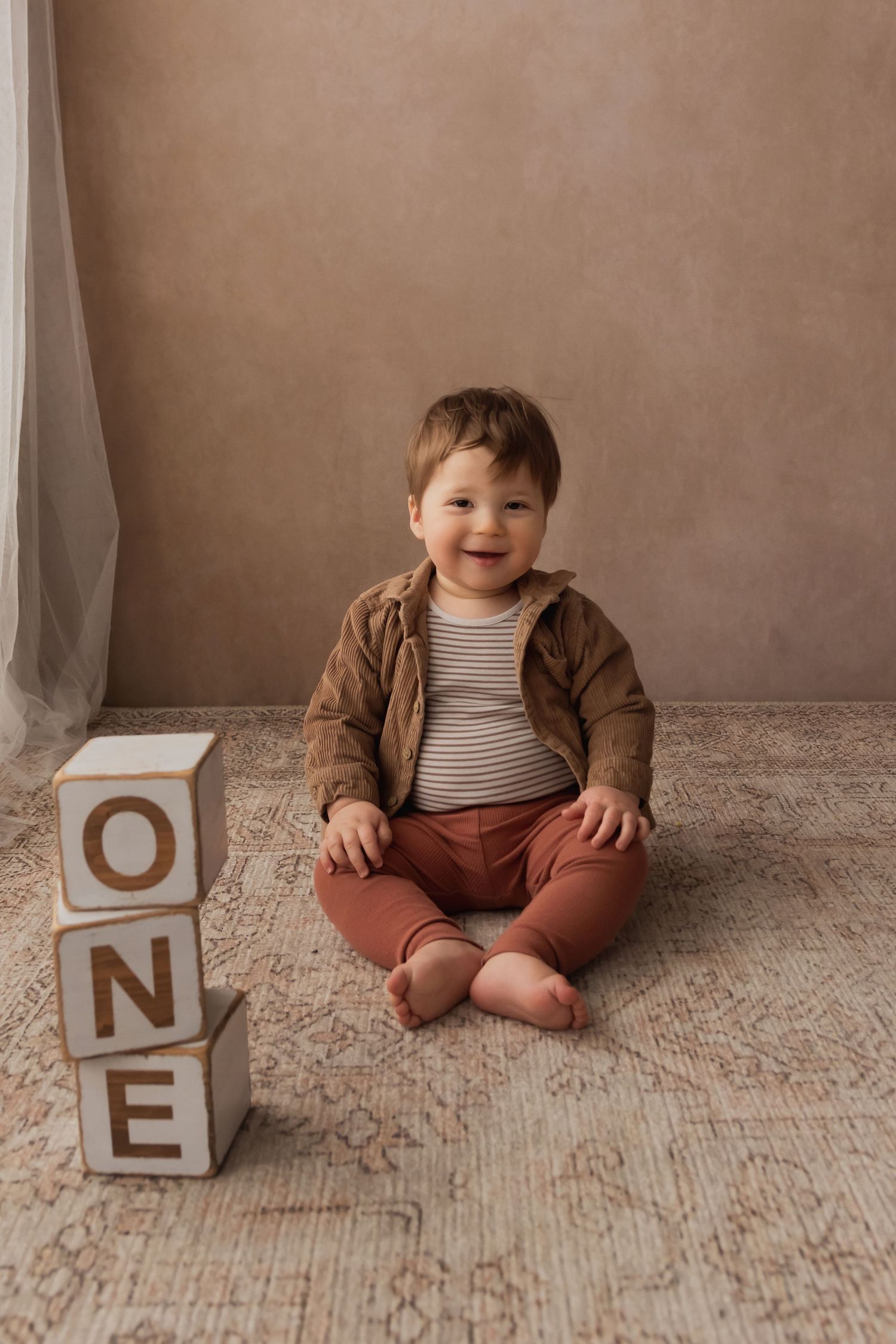 Cheerful one-year-old boy smiles beside stacked ONE letter blocks in warm brown and rust outfit.