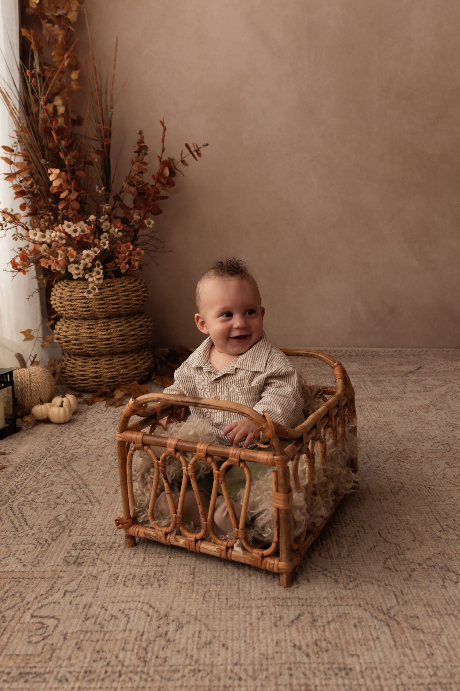 Smiling baby sitting in a rattan crib with autumn floral decor and warm beige backdrop in a cozy fall photo session.