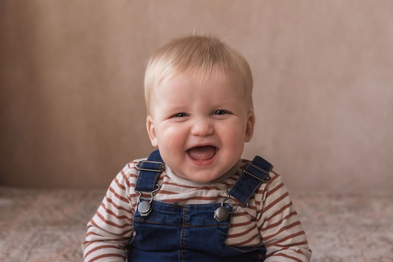 A cheerful blonde baby in striped shirt and denim overalls laughs joyfully during a photo session.