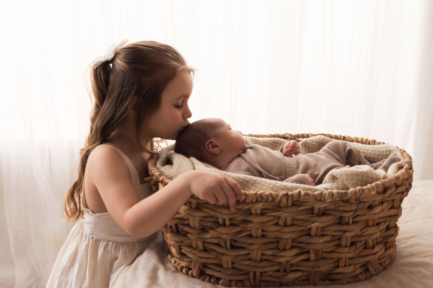 Young girl in a white dress gently kissing a sleeping newborn baby lying in a wicker basket.