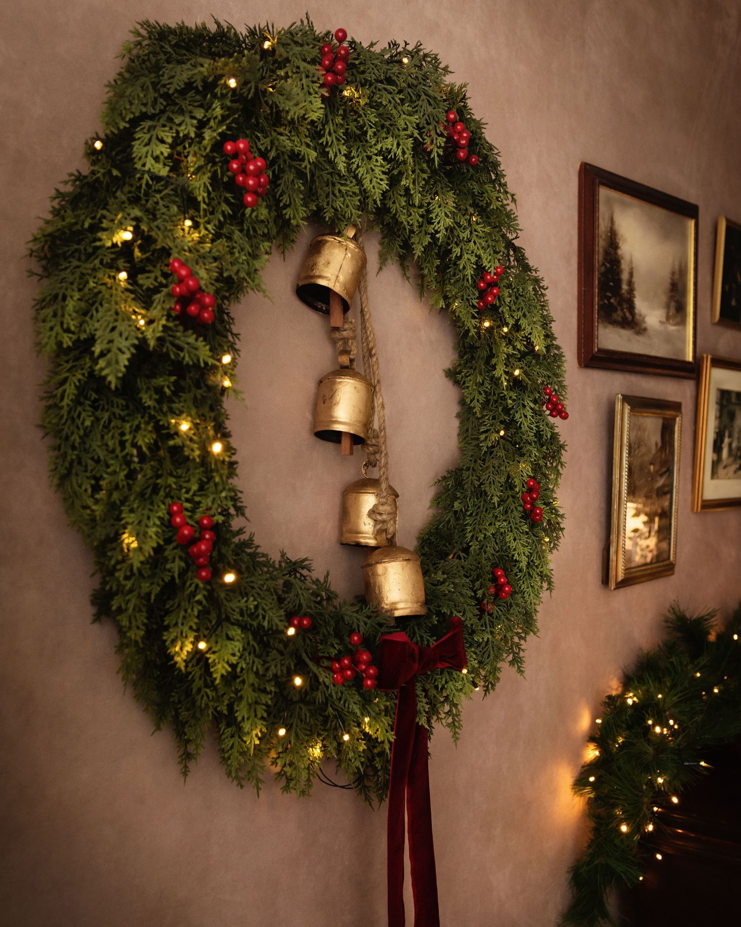 Large Christmas wreath with red berries, fairy lights, and gold bells with burgundy ribbon on a beige wall.