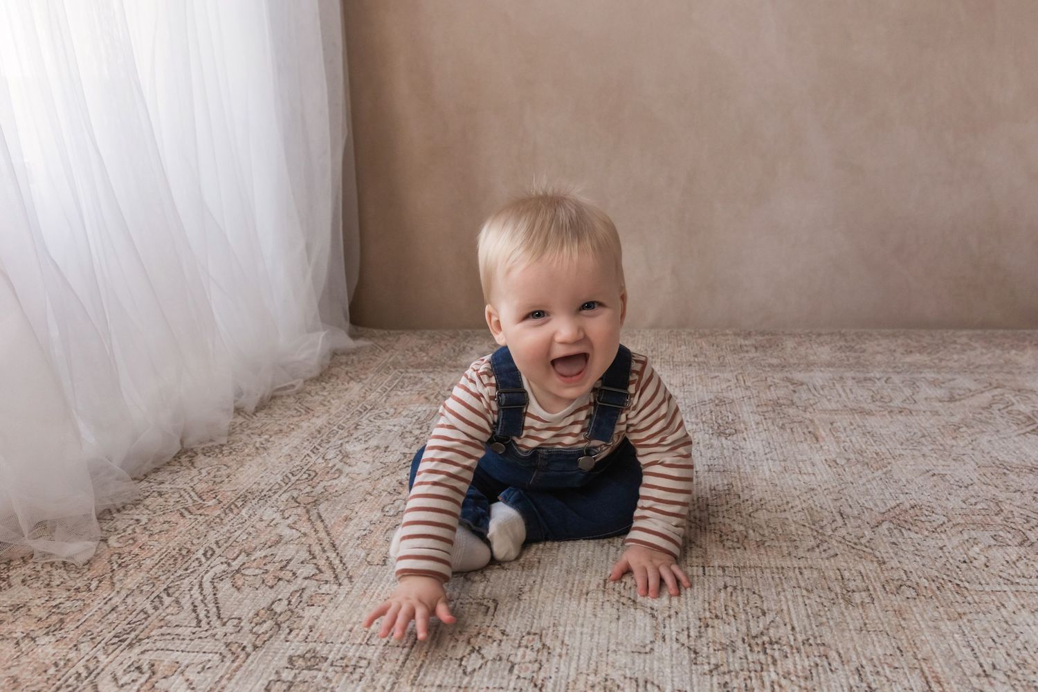 A cheerful blonde baby in denim overalls crawls on a patterned rug near a sheer white curtain.