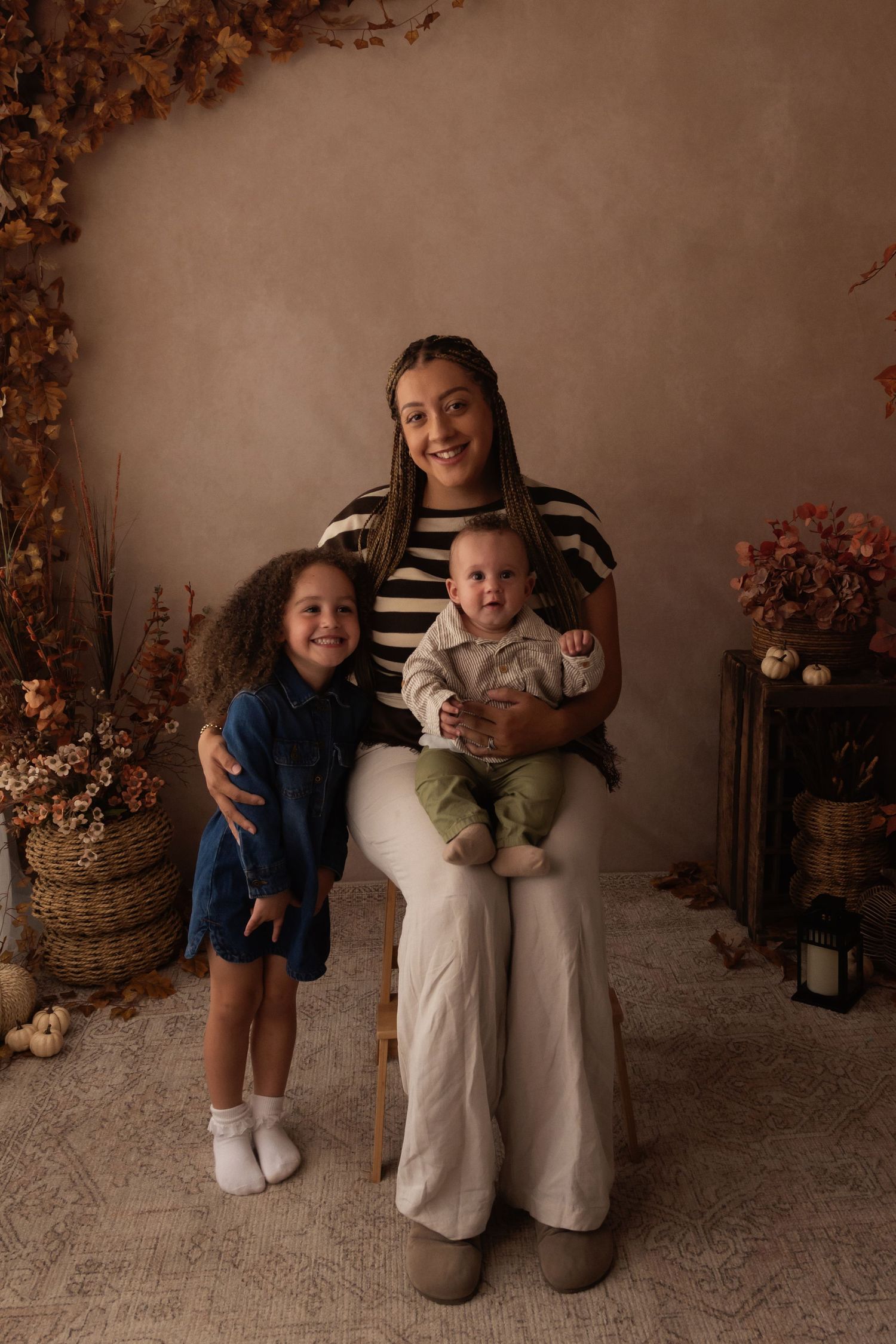 A woman sits holding a baby in green outfit while a young girl in blue dress stands beside her in fall-themed studio.