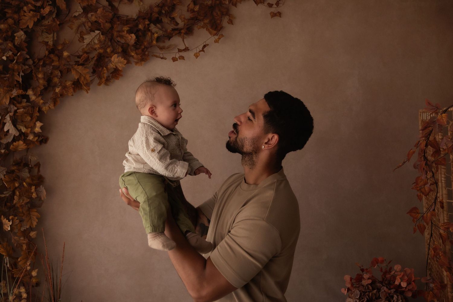 A bearded man holds a smiling baby up in the air against a warm autumn-themed studio backdrop.