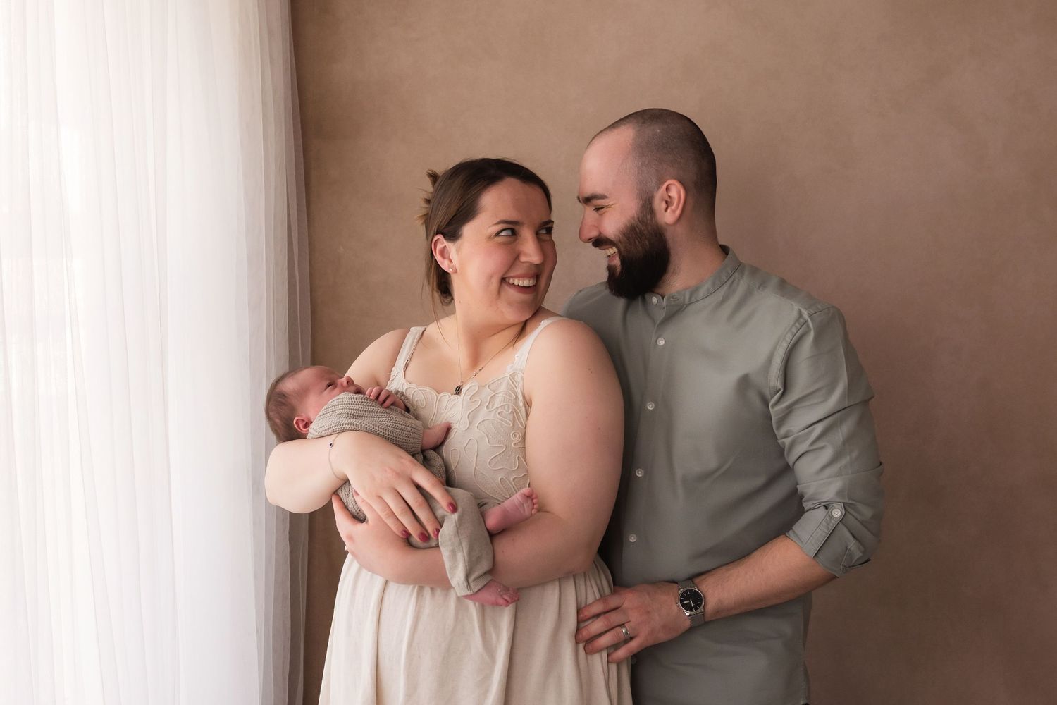 Smiling new parents holding newborn baby while standing near window in photo studio.