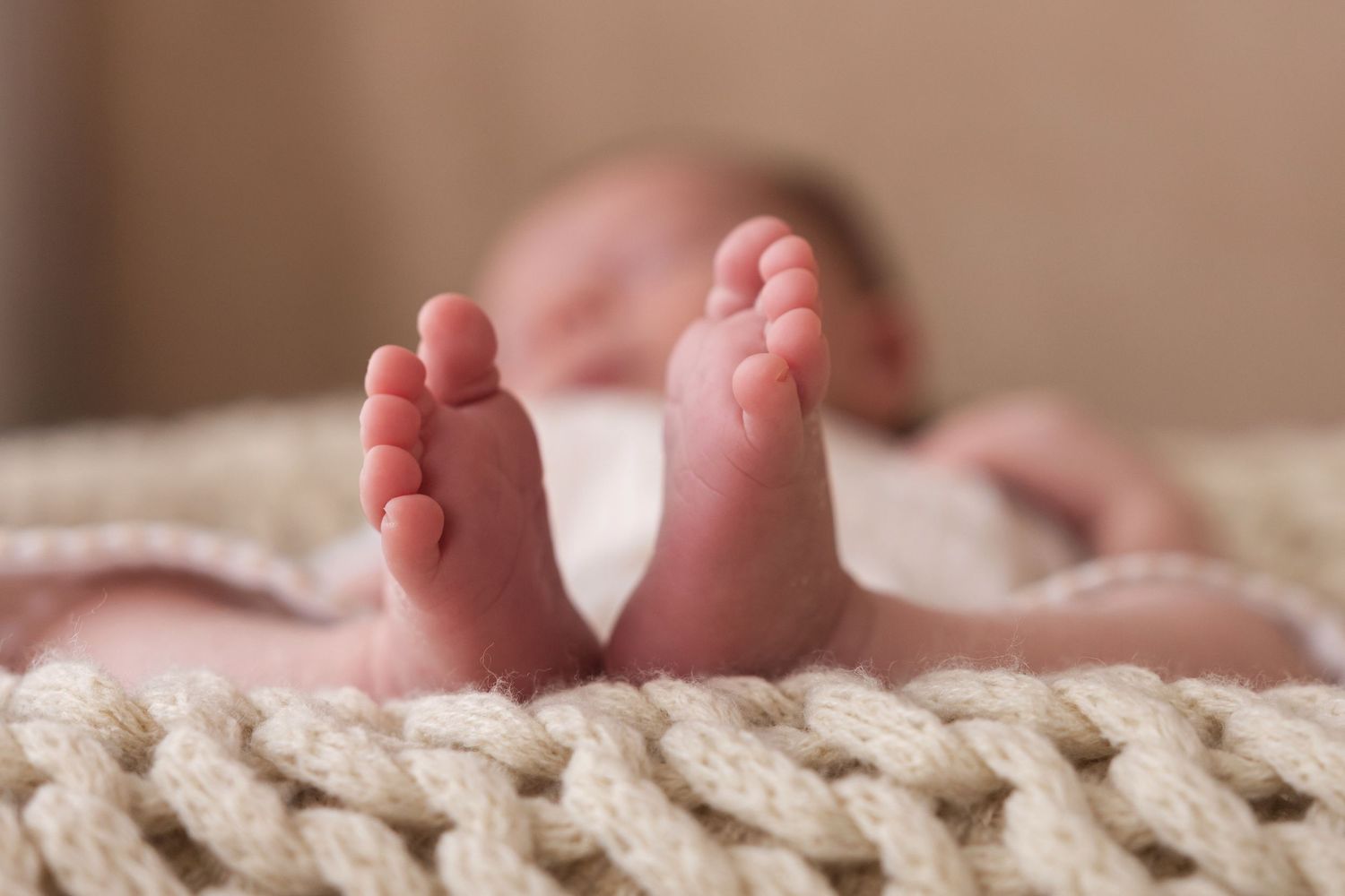Soft focus newborn baby feet in foreground with sleeping infant on cream knit blanket behind.