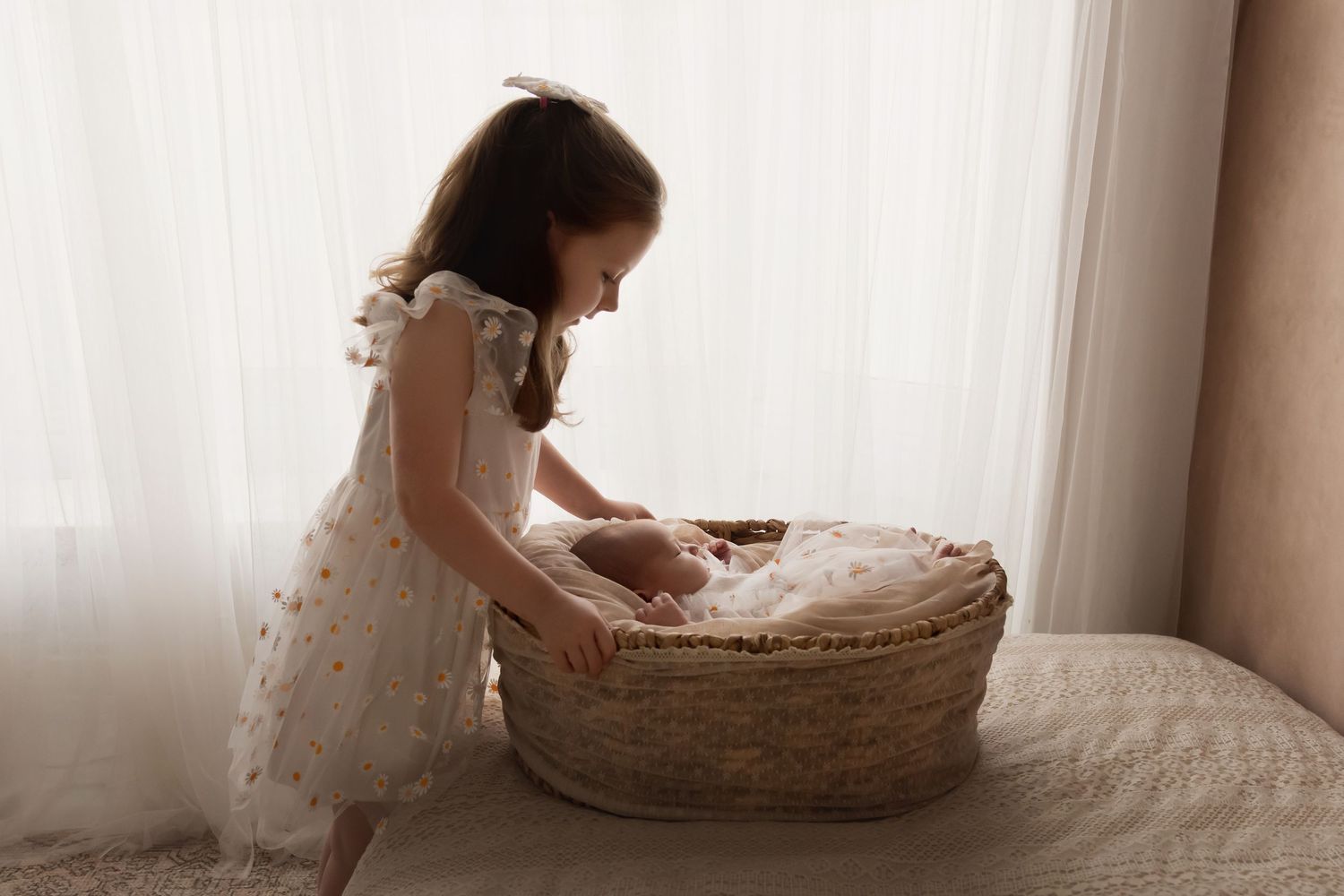 Young girl in white floral dress leaning over wicker Moses basket with newborn baby.